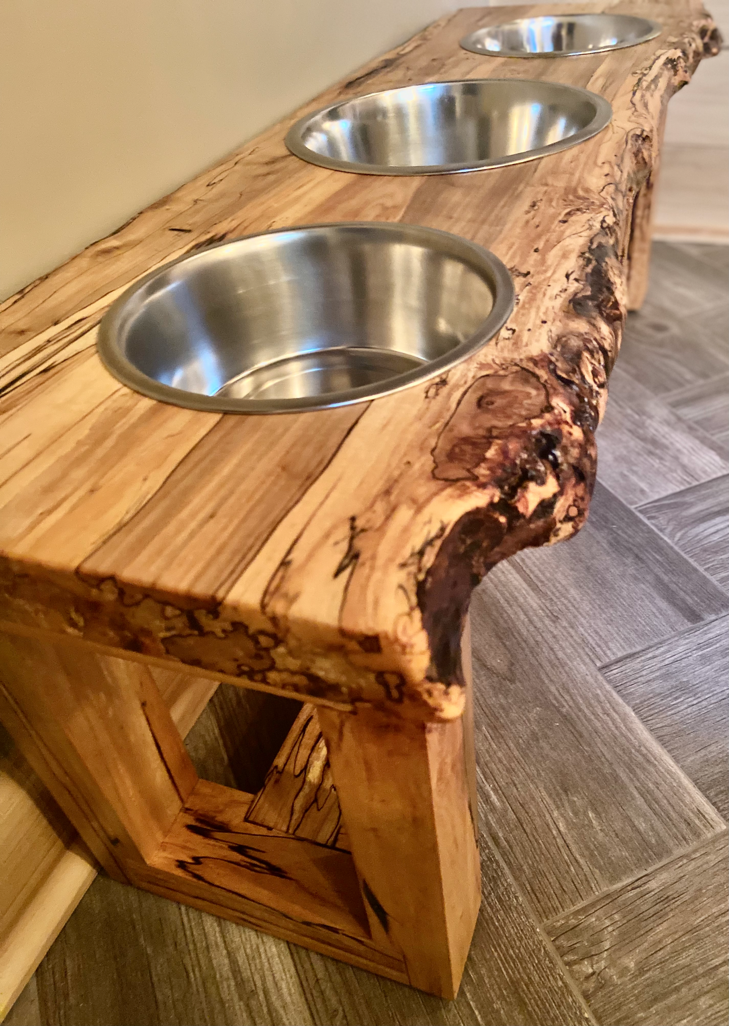 Live edge raised pet stand crafted from spalted maple or black walnut, featuring three stainless steel bowls for food and water. Elevated pet feeder, showcasing the natural wood grain and sturdy construction. Top-down view highlighting the placement of the two stainless steel bowls within the live edge wooden stand. Detailed close-up of the spalted maple’s unique wood grain patterns on the pet stand.
