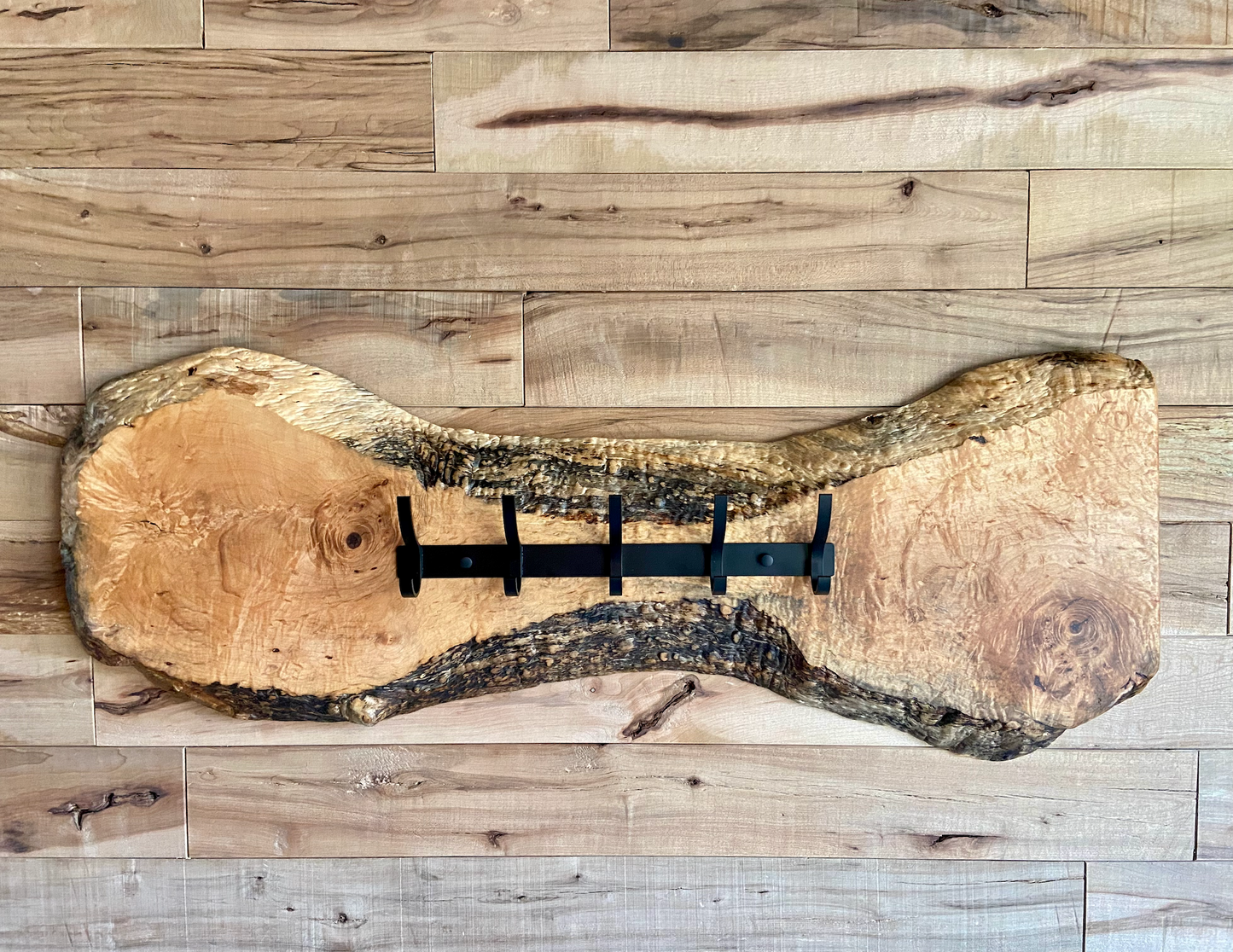 Live edge maple coat rack mounted on a wood-paneled wall, featuring 5 matte black metal hooks and natural wood grain patterns.