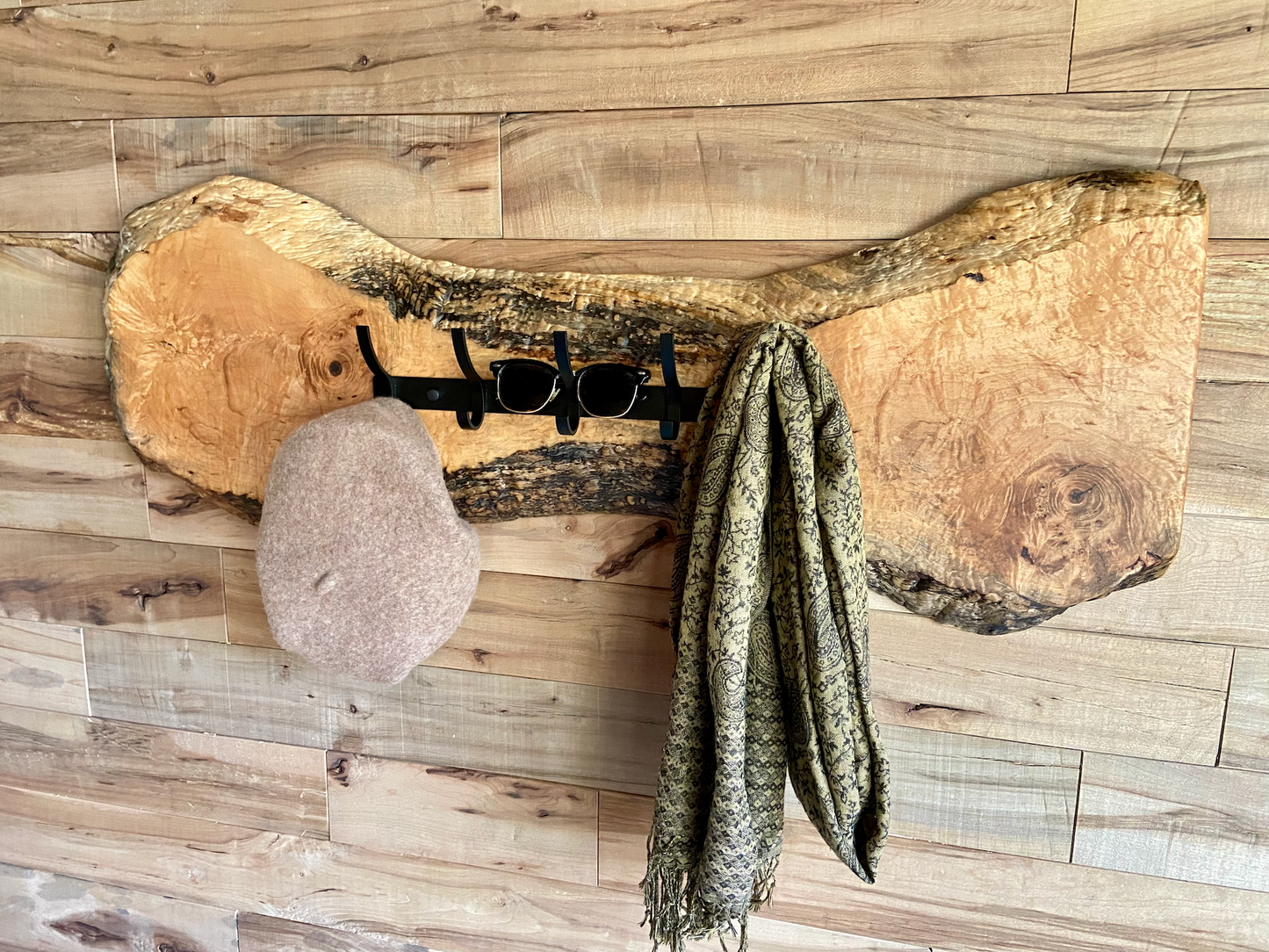 Live edge maple coat rack mounted on a wood-paneled wall with a hat and scarf hanging, featuring 5 matte black metal hooks and natural wood grain patterns.