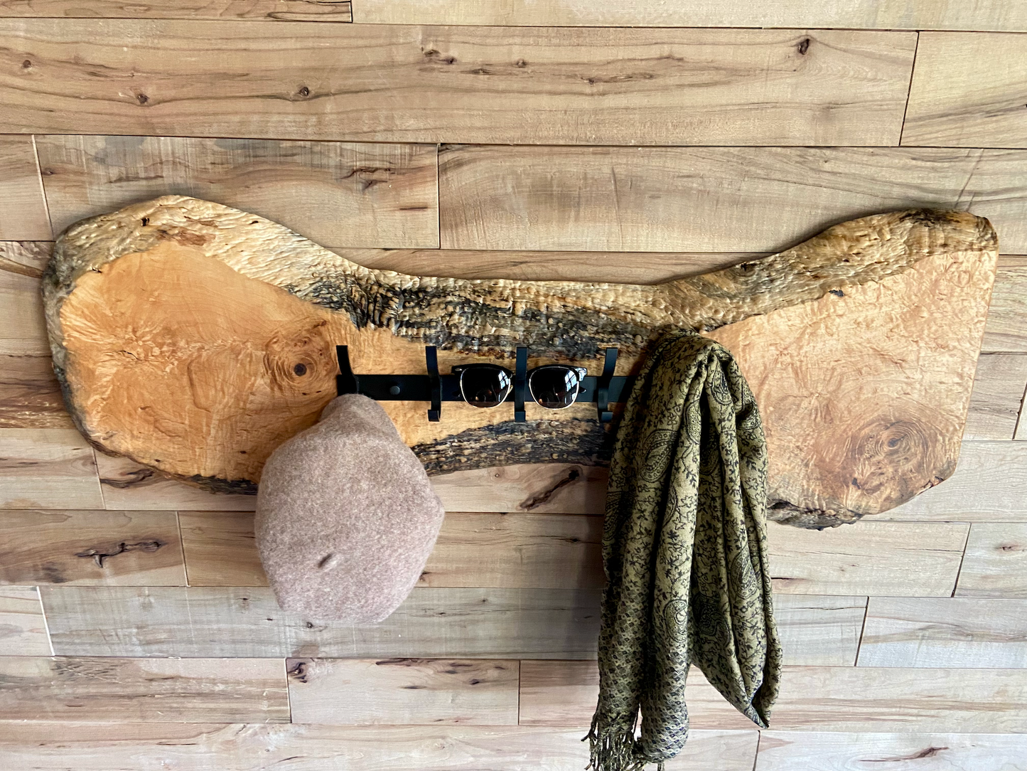Live edge maple coat rack mounted on a wood-paneled wall with a hat and scarf hanging, featuring 5 matte black metal hooks and natural wood grain patterns.