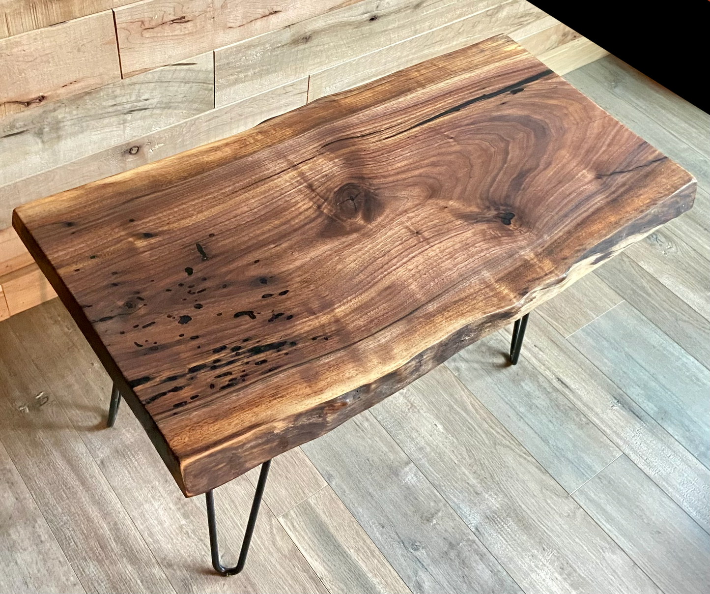 A handcrafted live edge walnut display table with black metal legs, placed against a couch. The live edge accent table showcases natural wood grain, knots, and organic edges, adding a rustic-modern touch to the space. A live edge walnut table styled with a candle, notebook, pen, a book, and a potted plant. The solid wood surface has a rich, natural finish, making it a perfect accent piece for an entryway or living space.