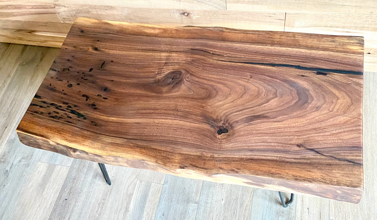 A handcrafted live edge walnut display table with black metal legs, placed against a couch. The live edge accent table showcases natural wood grain, knots, and organic edges, adding a rustic-modern touch to the space. A live edge walnut table styled with a candle, notebook, pen, a book, and a potted plant. The solid wood surface has a rich, natural finish, making it a perfect accent piece for an entryway or living space.