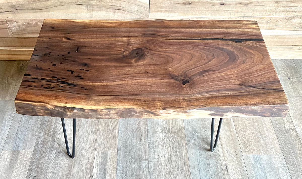 A handcrafted live edge walnut display table with black metal legs, placed against a couch. The live edge accent table showcases natural wood grain, knots, and organic edges, adding a rustic-modern touch to the space. A live edge walnut table styled with a candle, notebook, pen, a book, and a potted plant. The solid wood surface has a rich, natural finish, making it a perfect accent piece for an entryway or living space.