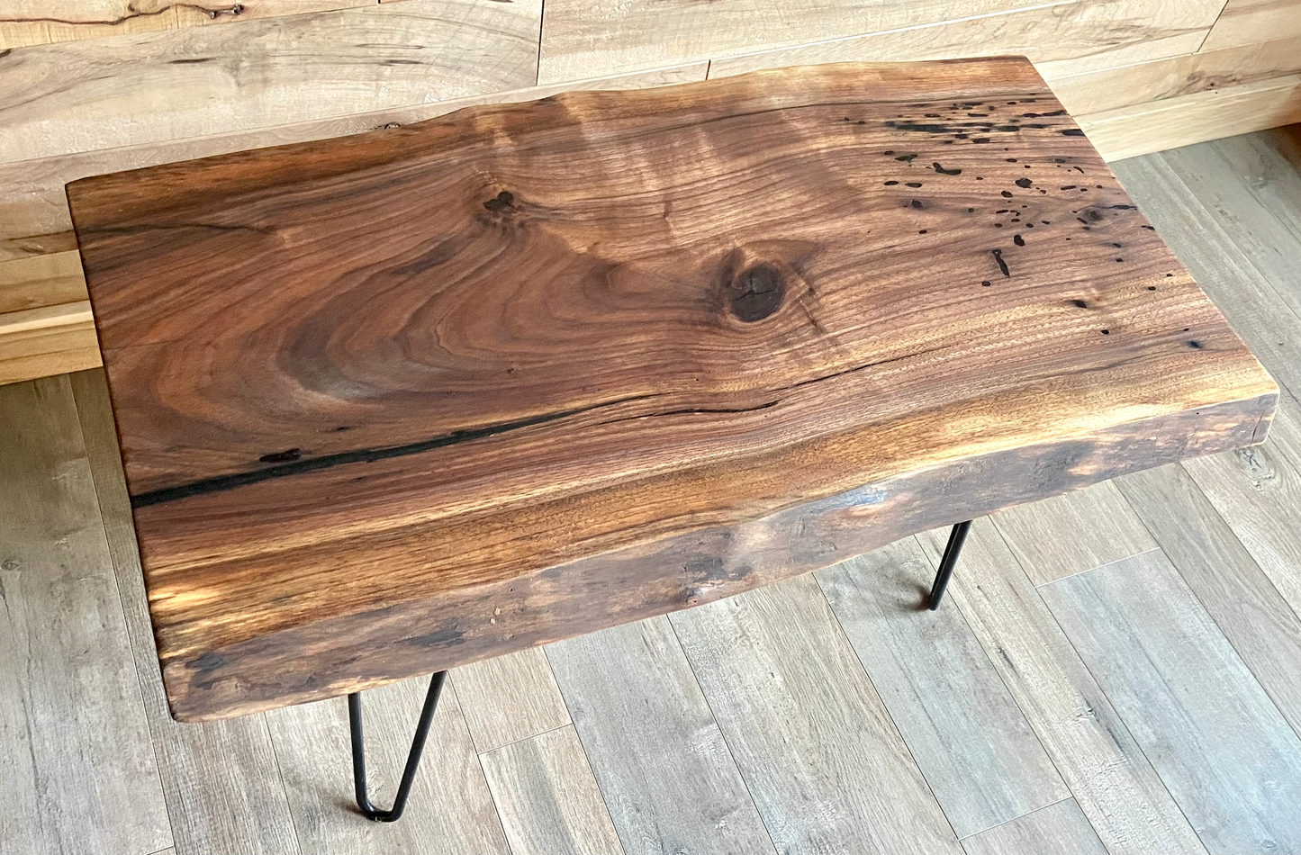 A handcrafted live edge walnut display table with black metal legs, placed against a couch. The live edge accent table showcases natural wood grain, knots, and organic edges, adding a rustic-modern touch to the space. A live edge walnut table styled with a candle, notebook, pen, a book, and a potted plant. The solid wood surface has a rich, natural finish, making it a perfect accent piece for an entryway or living space.