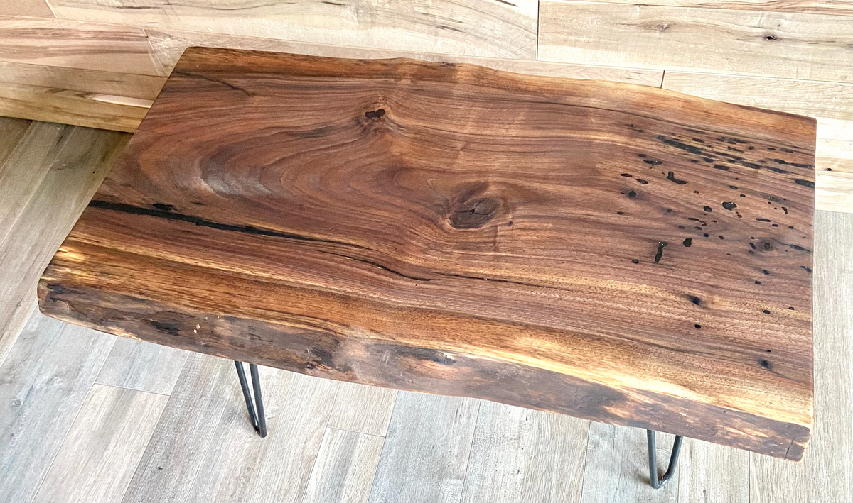 A handcrafted live edge walnut display table with black metal legs, placed against a couch. The live edge accent table showcases natural wood grain, knots, and organic edges, adding a rustic-modern touch to the space. A live edge walnut table styled with a candle, notebook, pen, a book, and a potted plant. The solid wood surface has a rich, natural finish, making it a perfect accent piece for an entryway or living space.