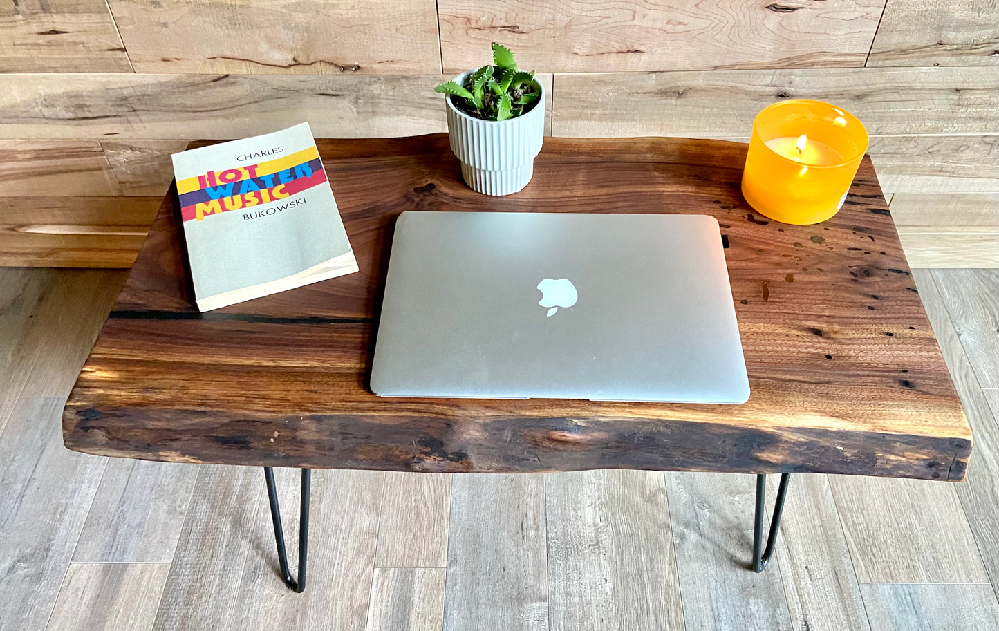 A handcrafted live edge walnut display table with black metal legs, placed against a couch. The live edge accent table showcases natural wood grain, knots, and organic edges, adding a rustic-modern touch to the space. A live edge walnut table styled with a candle, notebook, pen, a book, and a potted plant. The solid wood surface has a rich, natural finish, making it a perfect accent piece for an entryway or living space.