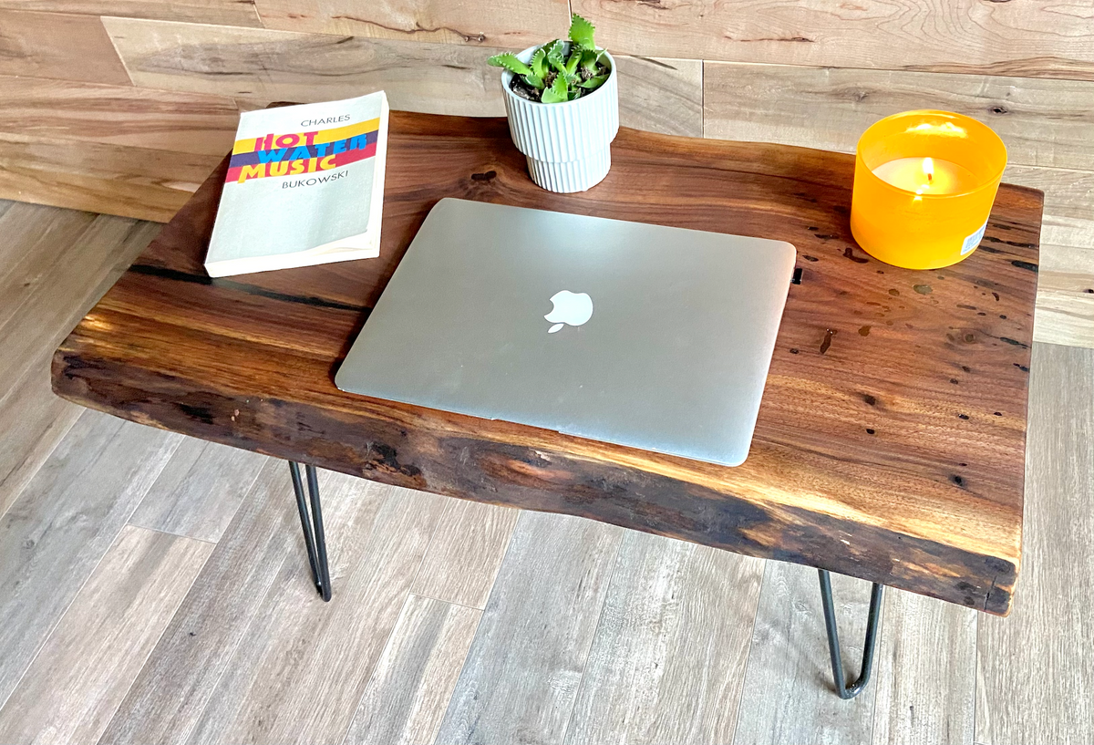 A handcrafted live edge walnut display table with black metal legs, placed against a couch. The live edge accent table showcases natural wood grain, knots, and organic edges, adding a rustic-modern touch to the space. A live edge walnut table styled with a candle, notebook, pen, a book, and a potted plant. The solid wood surface has a rich, natural finish, making it a perfect accent piece for an entryway or living space.