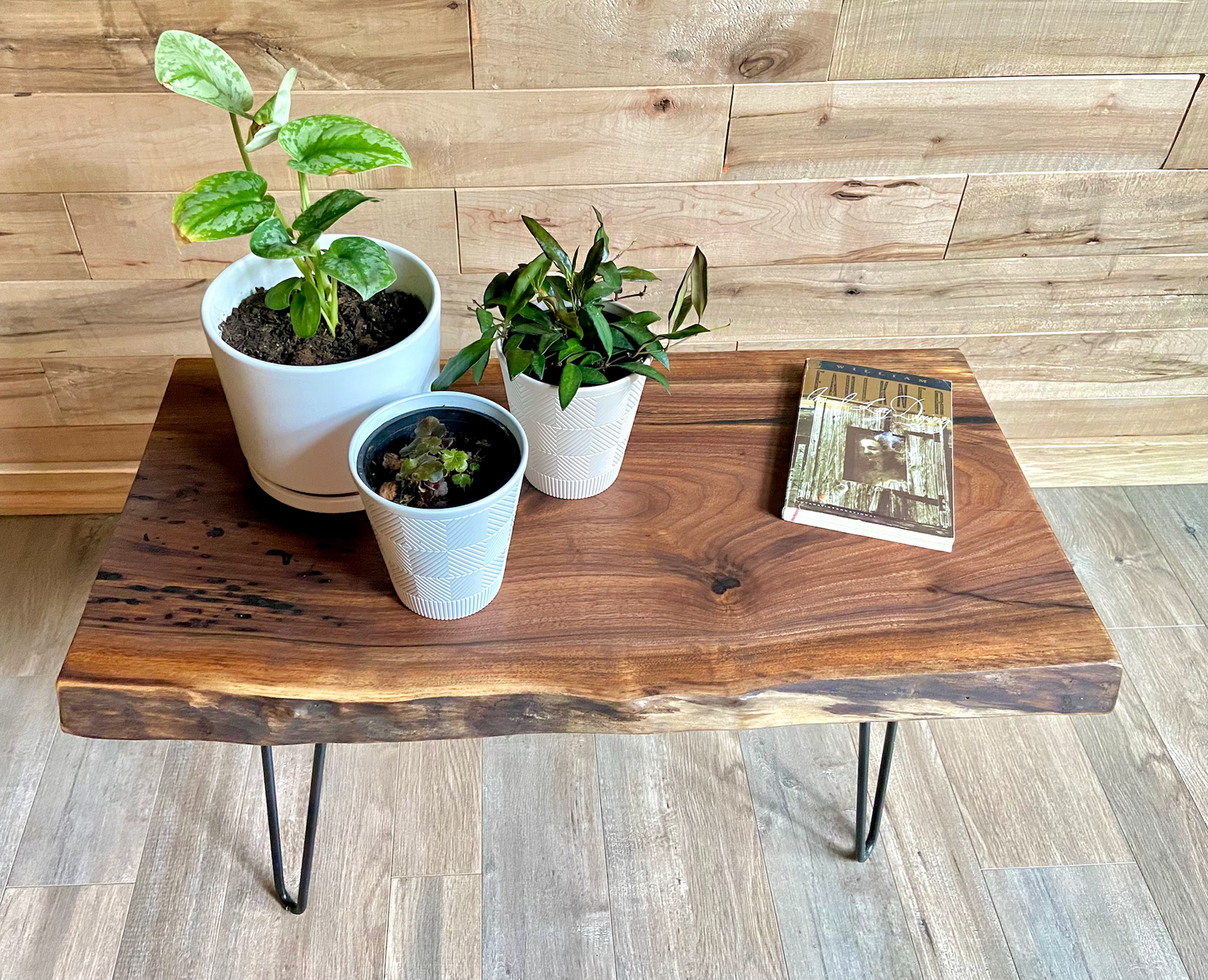 A handcrafted live edge walnut display table with black metal legs, placed against a couch. The live edge accent table showcases natural wood grain, knots, and organic edges, adding a rustic-modern touch to the space. A live edge walnut table styled with a candle, notebook, pen, a book, and a potted plant. The solid wood surface has a rich, natural finish, making it a perfect accent piece for an entryway or living space.