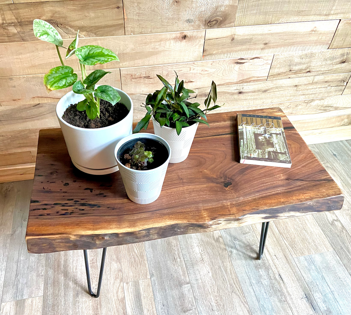 A handcrafted live edge walnut display table with black metal legs, placed against a couch. The live edge accent table showcases natural wood grain, knots, and organic edges, adding a rustic-modern touch to the space. A live edge walnut table styled with a candle, notebook, pen, a book, and a potted plant. The solid wood surface has a rich, natural finish, making it a perfect accent piece for an entryway or living space.
