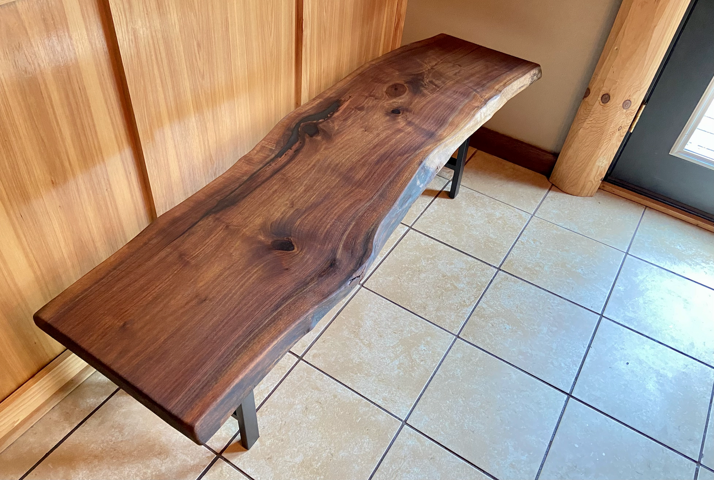 Live edge walnut bench with steel legs against a wood-paneled wall on tile flooring.