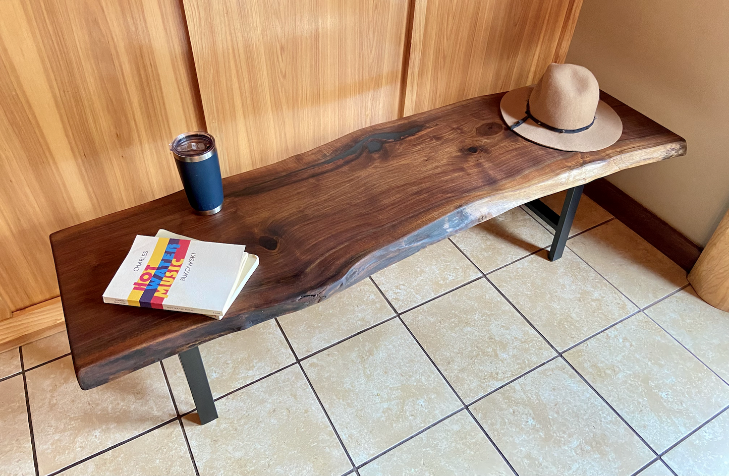 Live edge walnut bench with steel legs against a wood-paneled wall on tile flooring. Live edge walnut coffee table styled with everyday items, showcasing its versatility and natural wood grain.