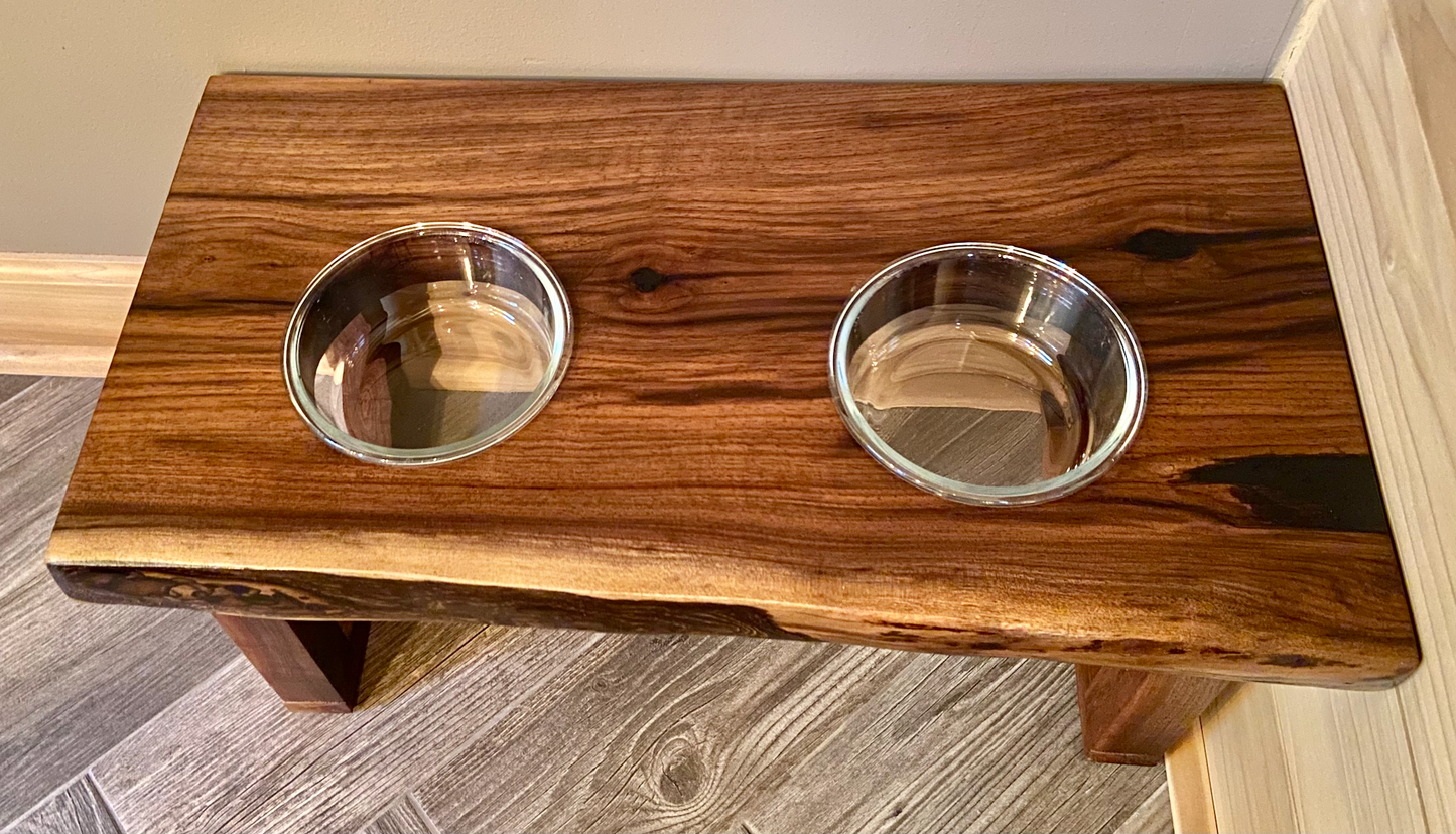 Live edge raised pet stand crafted from spalted maple or black walnut, featuring two stainless glass bowls for food and water. Elevated pet feeder, showcasing the natural wood grain and sturdy construction. Top-down view highlighting the placement of the two stainless steel bowls within the live edge wooden stand. Detailed close-up of the spalted maple’s unique wood grain patterns on the pet stand.