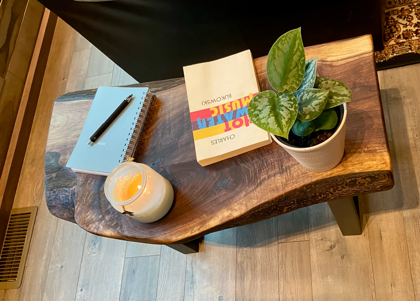 A handcrafted live edge walnut display table with black metal legs, placed against a couch. The live edge accent table showcases natural wood grain, knots, and organic edges, adding a rustic-modern touch to the space. A live edge walnut bench styled with a candle, notebook, pen, a book, and a potted plant. The solid wood surface has a rich, natural finish, making it a perfect accent piece for an entryway or living space.