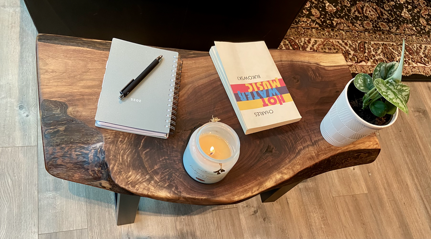 A handcrafted live edge walnut display table with black metal legs, placed against a couch. The live edge accent table showcases natural wood grain, knots, and organic edges, adding a rustic-modern touch to the space. A live edge walnut bench styled with a candle, notebook, pen, a book, and a potted plant. The solid wood surface has a rich, natural finish, making it a perfect accent piece for an entryway or living space.