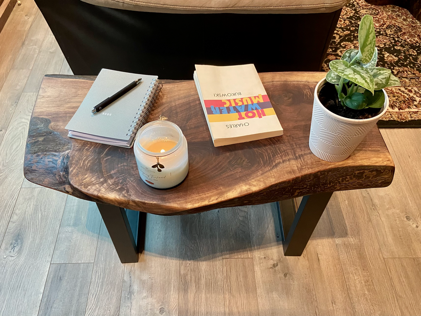 A handcrafted live edge walnut display table with black metal legs, placed against a couch. The live edge accent table showcases natural wood grain, knots, and organic edges, adding a rustic-modern touch to the space. A live edge walnut bench styled with a candle, notebook, pen, a book, and a potted plant. The solid wood surface has a rich, natural finish, making it a perfect accent piece for an entryway or living space.