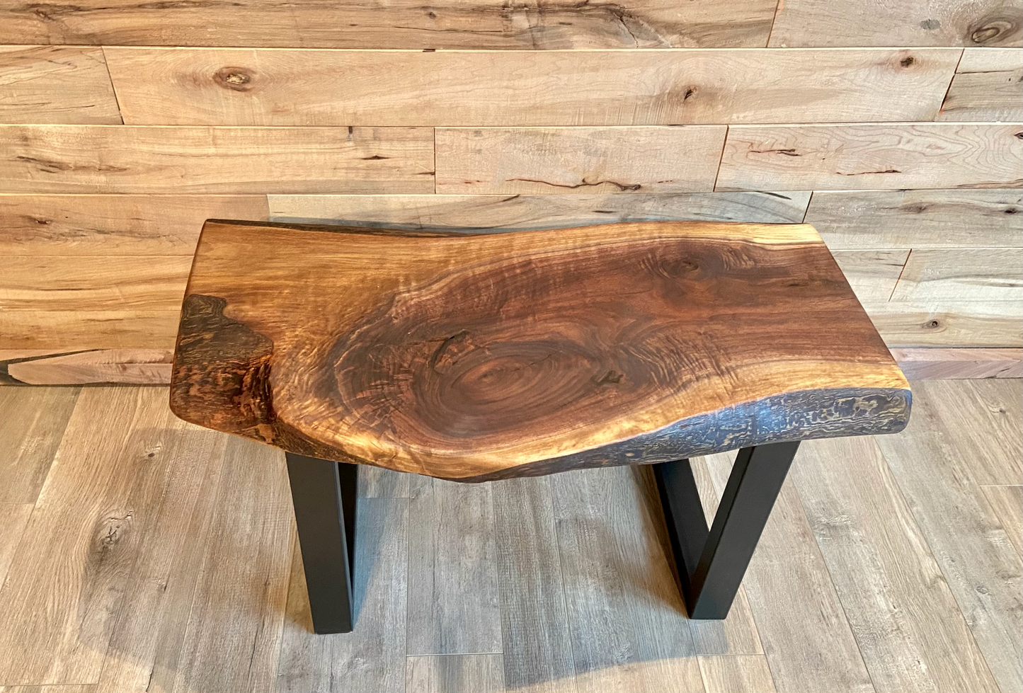 A handcrafted live edge walnut display table with black metal legs, placed against a wooden accent wall. The live edge accent table showcases natural wood grain, knots, and organic edges, adding a rustic-modern touch to the space. The solid wood surface has a rich, natural finish, making it a perfect accent piece for an entryway or living space.