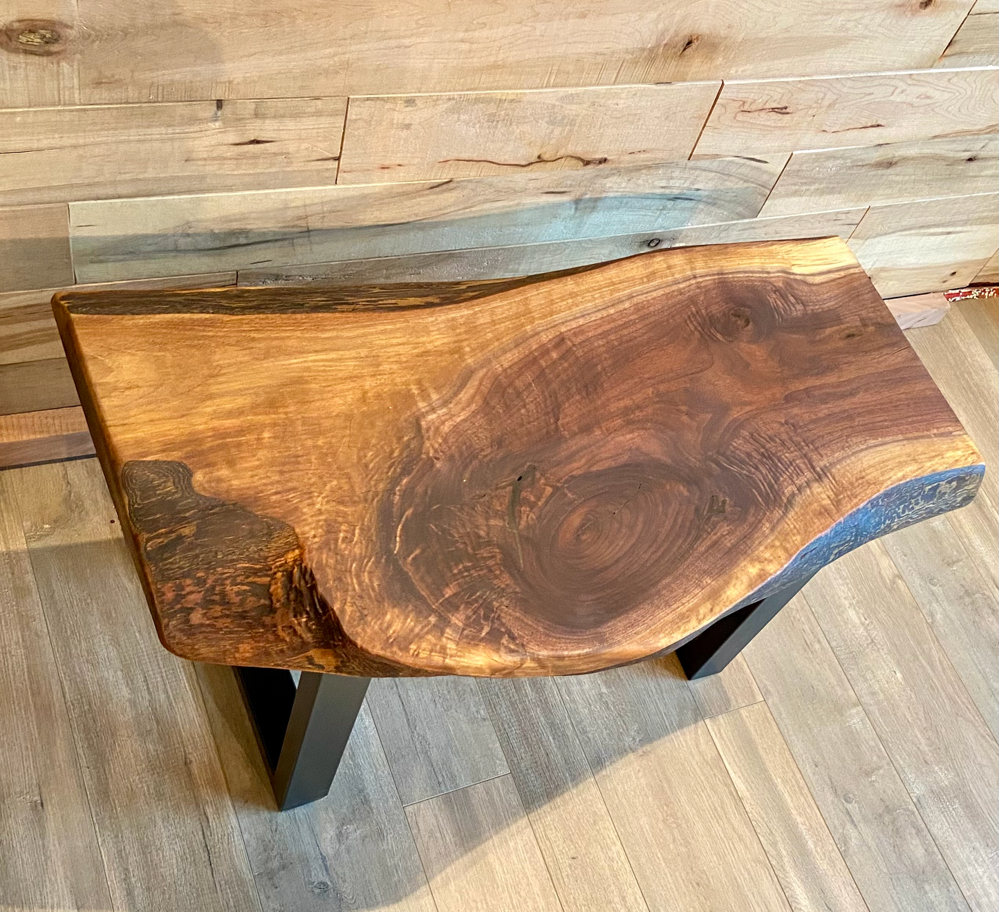 A handcrafted live edge walnut display table with black metal legs, placed against a wooden accent wall. The live edge accent table showcases natural wood grain, knots, and organic edges, adding a rustic-modern touch to the space. The solid wood surface has a rich, natural finish, making it a perfect accent piece for an entryway or living space.