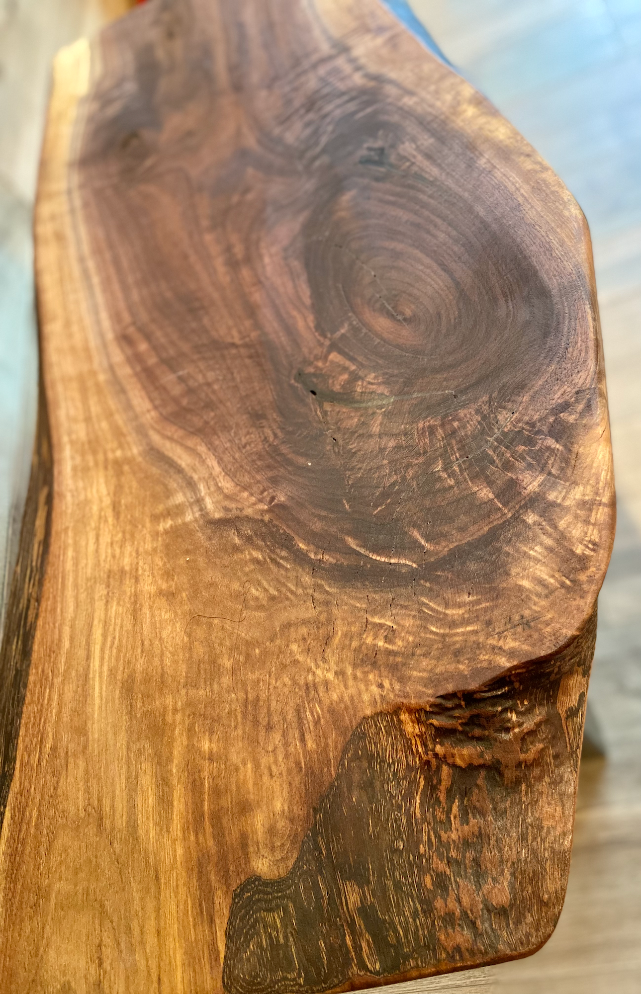 A handcrafted live edge walnut display table with black metal legs, placed against a wooden accent wall. The live edge accent table showcases natural wood grain, knots, and organic edges, adding a rustic-modern touch to the space. The solid wood surface has a rich, natural finish, making it a perfect accent piece for an entryway or living space.