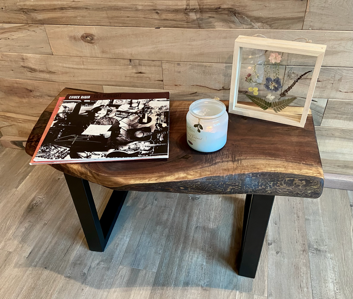 A handcrafted live edge walnut display table with black metal legs, placed against a wooden accent wall. The live edge accent table showcases natural wood grain, knots, and organic edges, adding a rustic-modern touch to the space. A live edge walnut table styled with a candle, notebook, pen, a book, and a potted plant. The solid wood surface has a rich, natural finish, making it a perfect accent piece for an entryway or living space.
