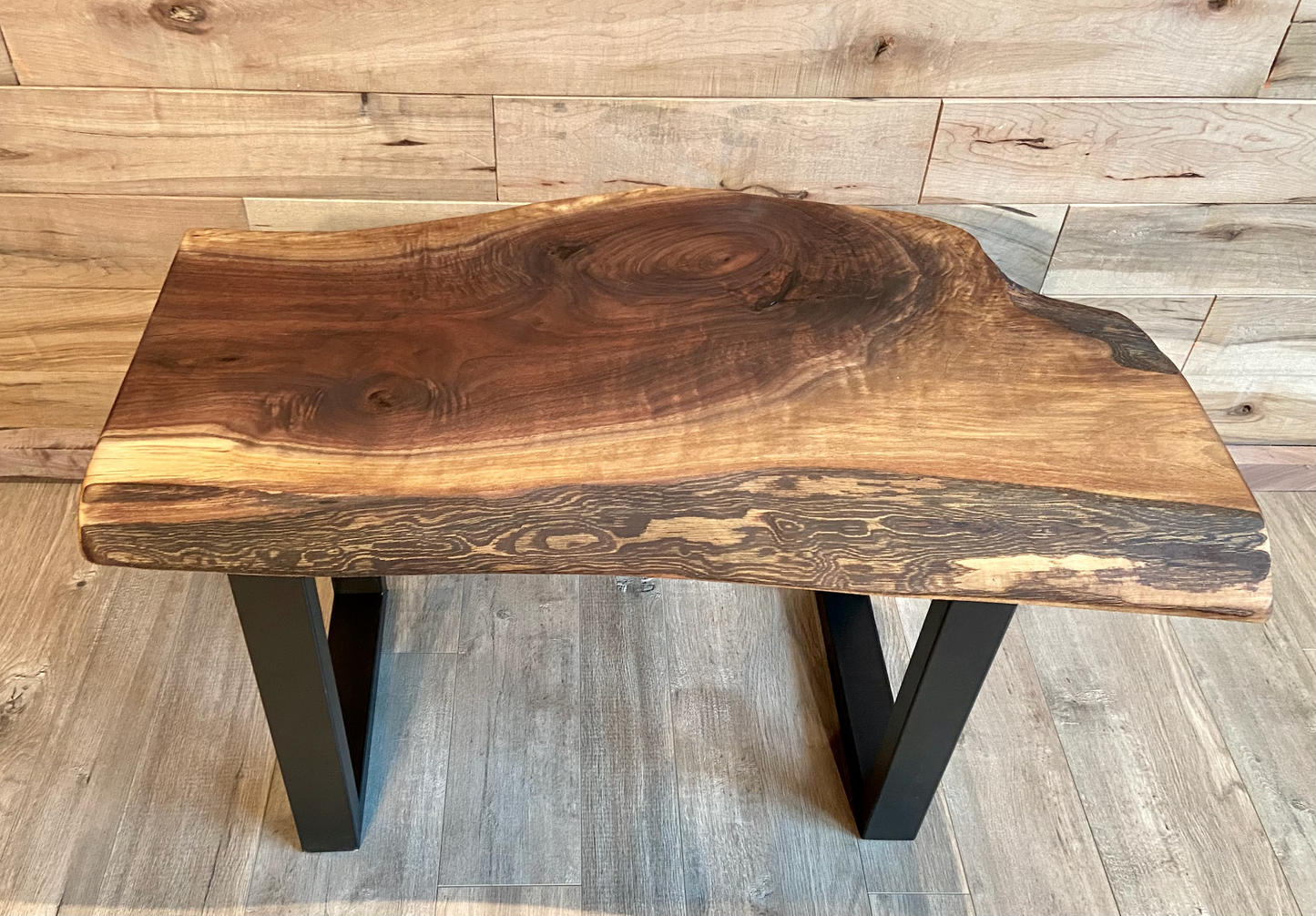 A handcrafted live edge walnut display table with black metal legs, placed against a wooden accent wall. The live edge accent table showcases natural wood grain, knots, and organic edges, adding a rustic-modern touch to the space. The solid wood surface has a rich, natural finish, making it a perfect accent piece for an entryway or living space.