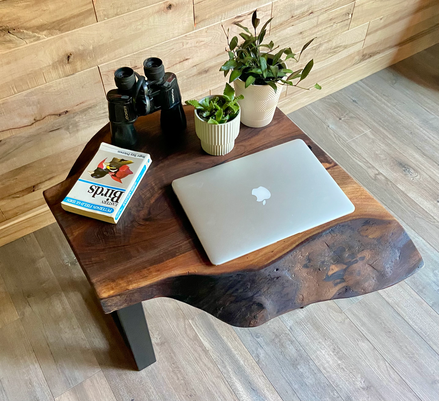 Live edge walnut table with laptop, book, binoculars, and plants against a wooden wall.