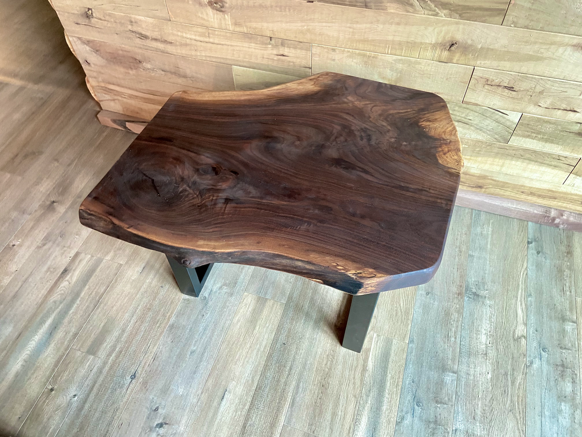 A handcrafted live edge walnut display table with black metal legs, placed against a wooden accent wall. The live edge accent table showcases natural wood grain, knots, and organic edges, adding a rustic-modern touch to the space. The solid wood surface has a rich, natural finish, making it a perfect accent piece for an entryway or living space.