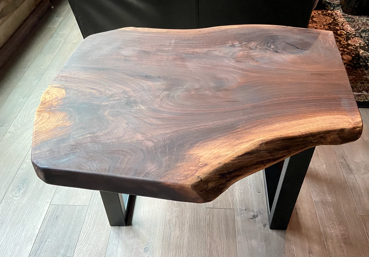 A handcrafted live edge walnut display table with black metal legs, placed against a couch. The live edge accent table showcases natural wood grain, knots, and organic edges, adding a rustic-modern touch to the space. The solid wood surface has a rich, natural finish, making it a perfect accent piece for an entryway or living space.