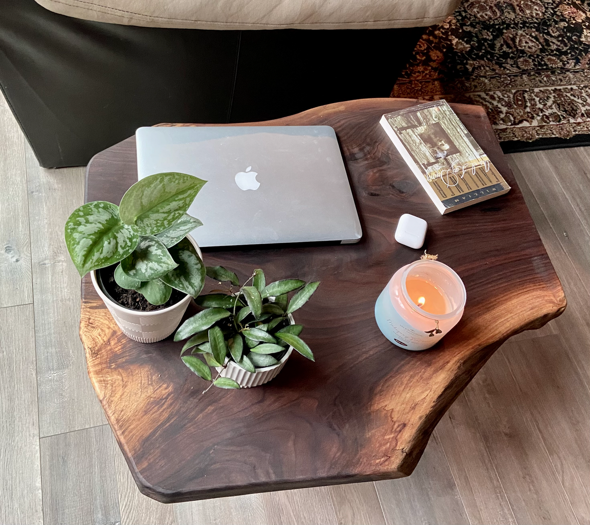 A handcrafted live edge walnut display table with black metal legs, placed against a couch. The live edge accenttable showcases natural wood grain, knots, and organic edges, adding a rustic-modern touch to the space. A live edge walnut bench styled with a candle, notebook, pen, a book, and a potted plant. The solid wood surface has a rich, natural finish, making it a perfect accent piece for an entryway or living space.