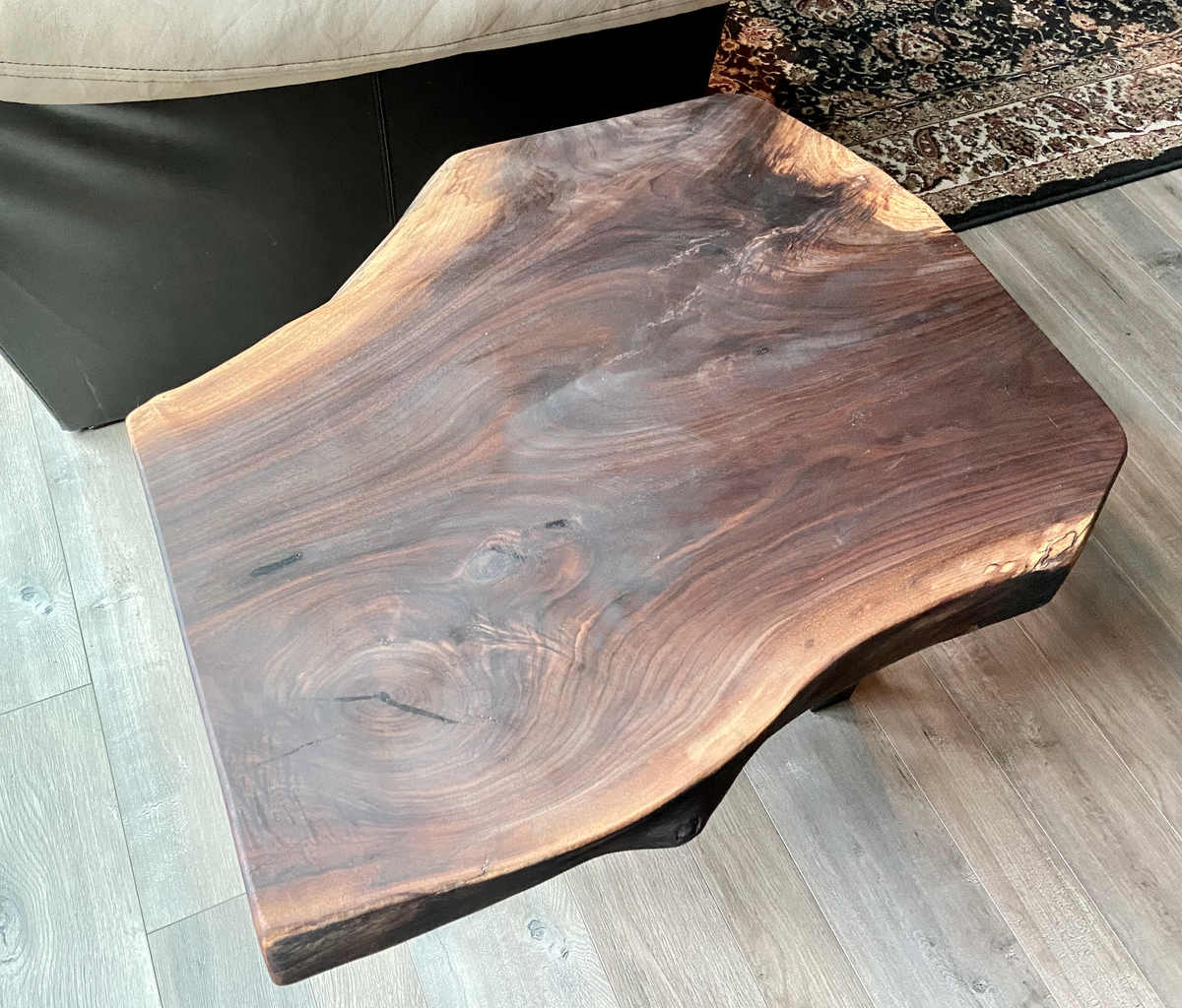 A handcrafted live edge walnut display table with black metal legs, placed against a couch. The live edge accent table showcases natural wood grain, knots, and organic edges, adding a rustic-modern touch to the space. The solid wood surface has a rich, natural finish, making it a perfect accent piece for an entryway or living space.