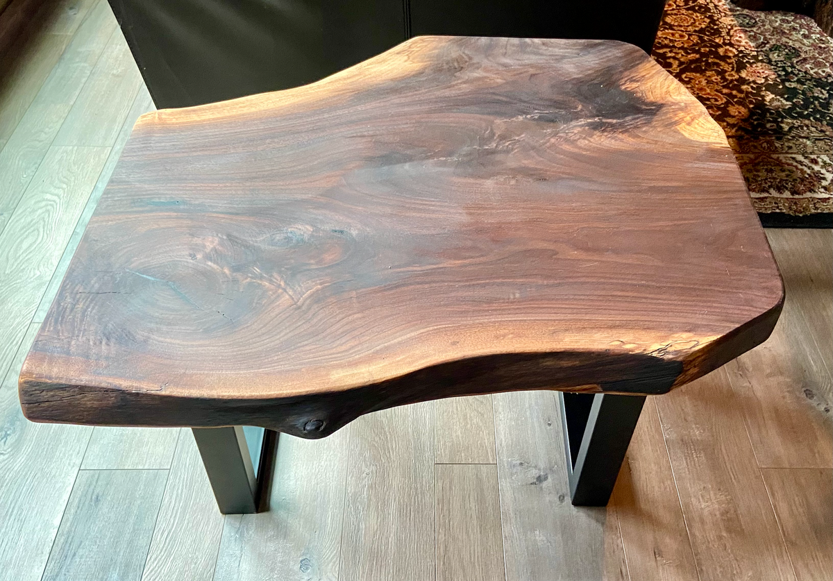 A handcrafted live edge walnut display table with black metal legs, placed against a couch. The live edge accent table showcases natural wood grain, knots, and organic edges, adding a rustic-modern touch to the space. The solid wood surface has a rich, natural finish, making it a perfect accent piece for an entryway or living space.