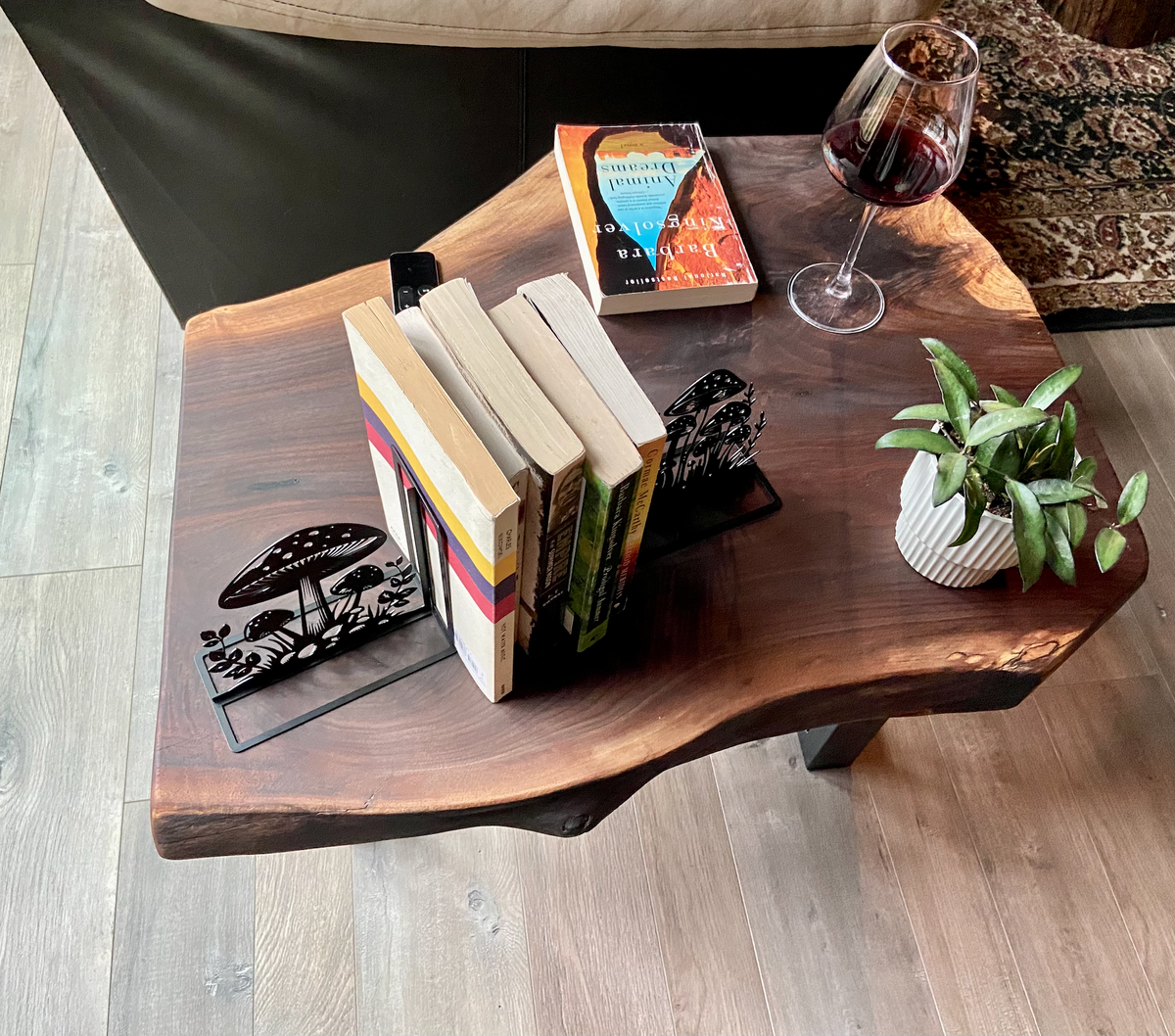 A handcrafted live edge walnut display table with black metal legs, placed against a couch. The live edge accenttable showcases natural wood grain, knots, and organic edges, adding a rustic-modern touch to the space. A live edge walnut bench styled with a candle, notebook, pen, a book, and a potted plant. The solid wood surface has a rich, natural finish, making it a perfect accent piece for an entryway or living space.