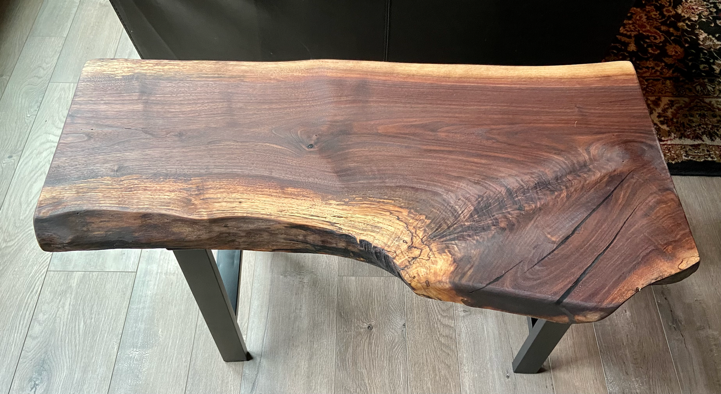 A handcrafted live edge walnut display table with black metal legs, placed against a couch. The live edge accent table showcases natural wood grain, knots, and organic edges, adding a rustic-modern touch to the space. The solid wood surface has a rich, natural finish, making it a perfect accent piece for an entryway or living space.
