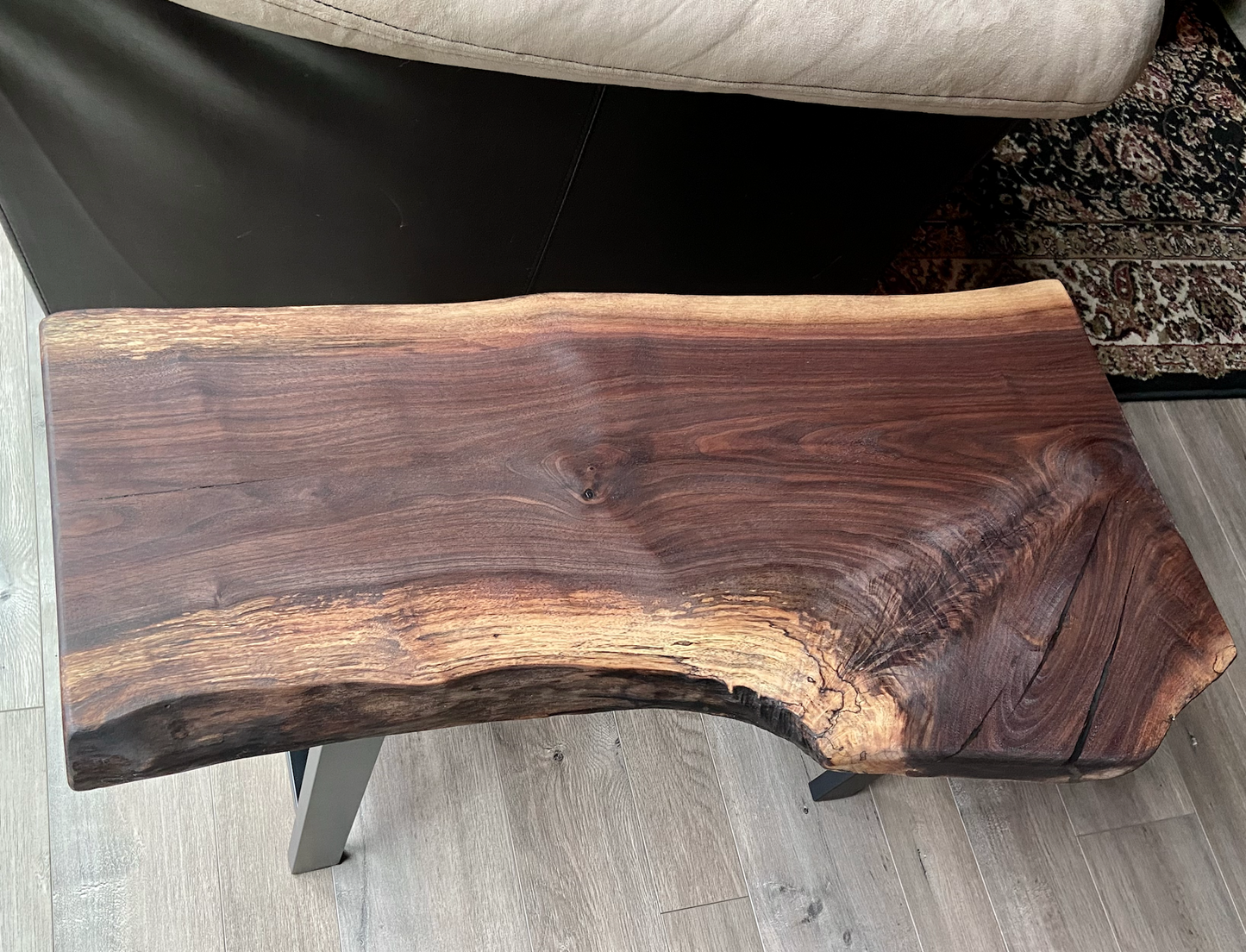 A handcrafted live edge walnut display table with black metal legs, placed against a couch. The live edge accenttable showcases natural wood grain, knots, and organic edges, adding a rustic-modern touch to the space. The solid wood surface has a rich, natural finish, making it a perfect accent piece for an entryway or living space.