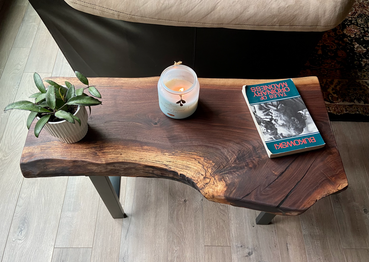 A handcrafted live edge walnut display table with black metal legs, placed against a couch. The live edge accenttable showcases natural wood grain, knots, and organic edges, adding a rustic-modern touch to the space. A live edge walnut bench styled with a candle, notebook, pen, a book, and a potted plant. The solid wood surface has a rich, natural finish, making it a perfect accent piece for an entryway or living space.