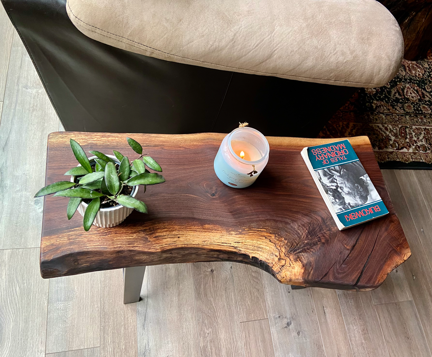 A handcrafted live edge walnut display table with black metal legs, placed against a couch. The live edge accenttable showcases natural wood grain, knots, and organic edges, adding a rustic-modern touch to the space. A live edge walnut bench styled with a candle, notebook, pen, a book, and a potted plant. The solid wood surface has a rich, natural finish, making it a perfect accent piece for an entryway or living space.
