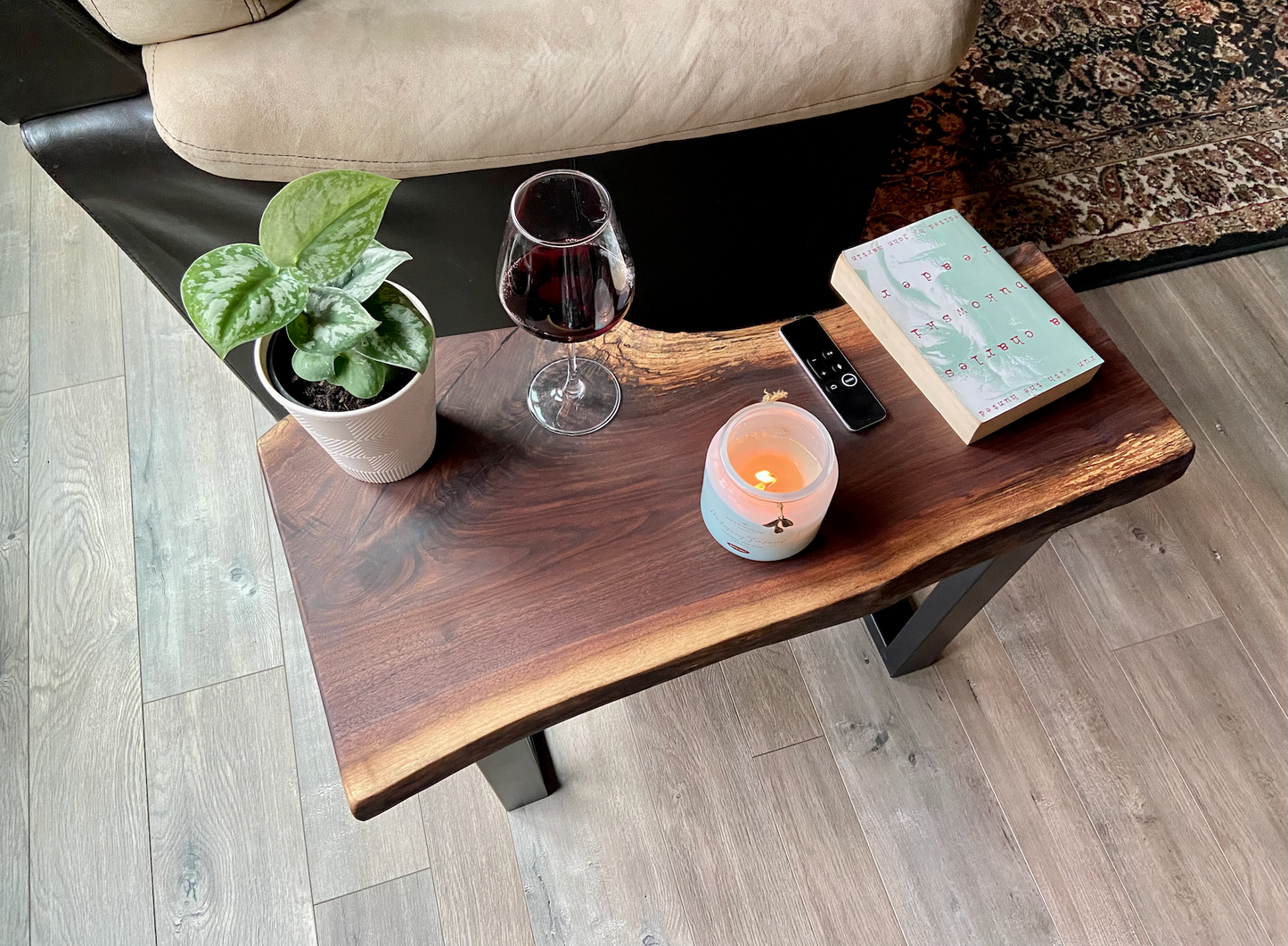 A handcrafted live edge walnut display table with black metal legs, placed against a couch. The live edge accenttable showcases natural wood grain, knots, and organic edges, adding a rustic-modern touch to the space. A live edge walnut bench styled with a candle, notebook, pen, a book, and a potted plant. The solid wood surface has a rich, natural finish, making it a perfect accent piece for an entryway or living space.
