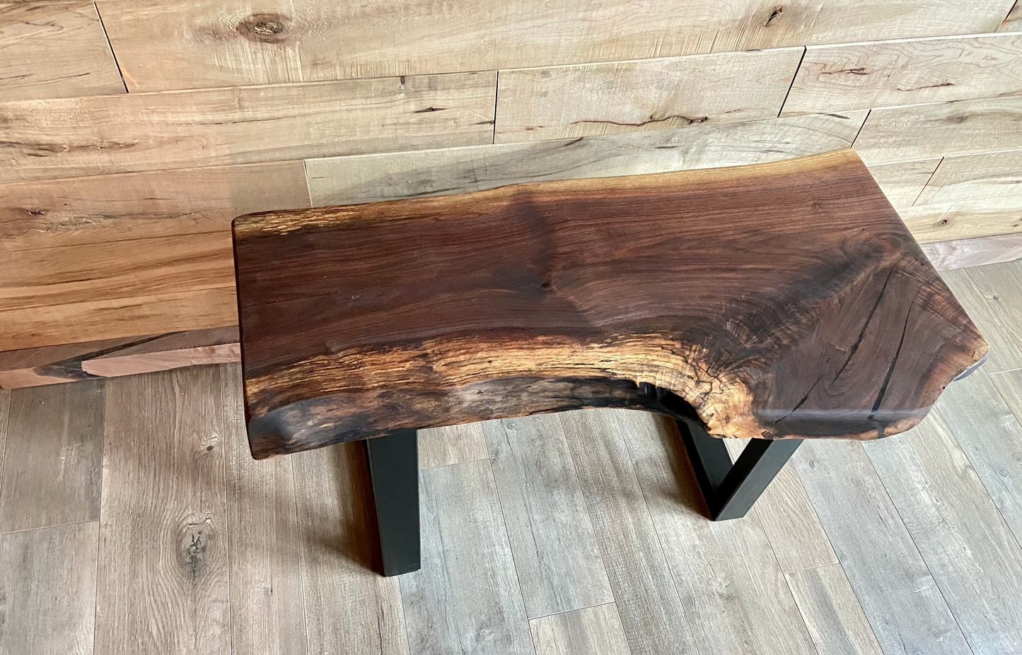 A handcrafted live edge walnut display table with black metal legs, placed against a wooden accent wall. The live edge accent table showcases natural wood grain, knots, and organic edges, adding a rustic-modern touch to the space. The solid wood surface has a rich, natural finish, making it a perfect accent piece for an entryway or living space.