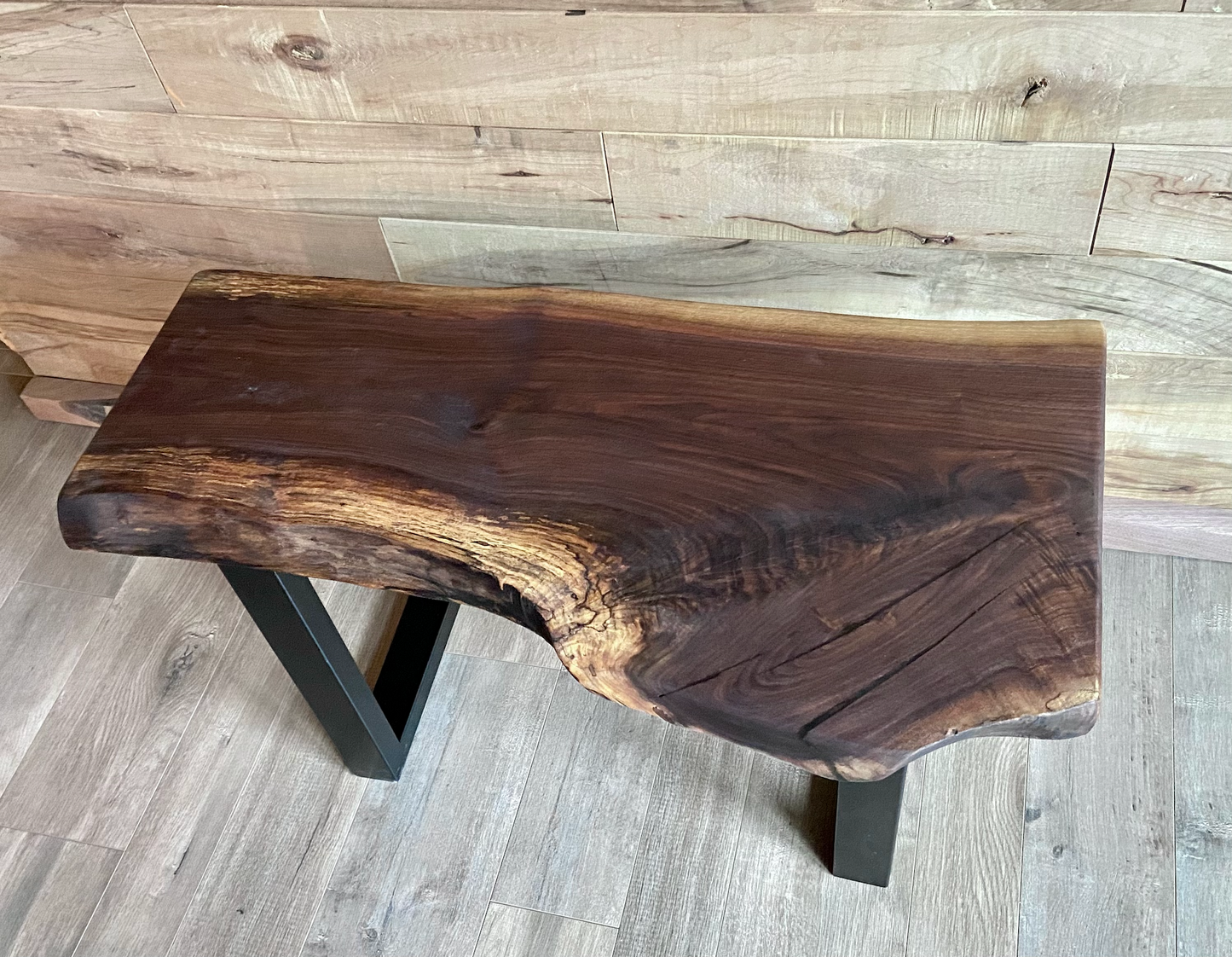 A handcrafted live edge walnut display table with black metal legs, placed against a wooden accent wall. The live edge accenttable showcases natural wood grain, knots, and organic edges, adding a rustic-modern touch to the space. The solid wood surface has a rich, natural finish, making it a perfect accent piece for an entryway or living space.