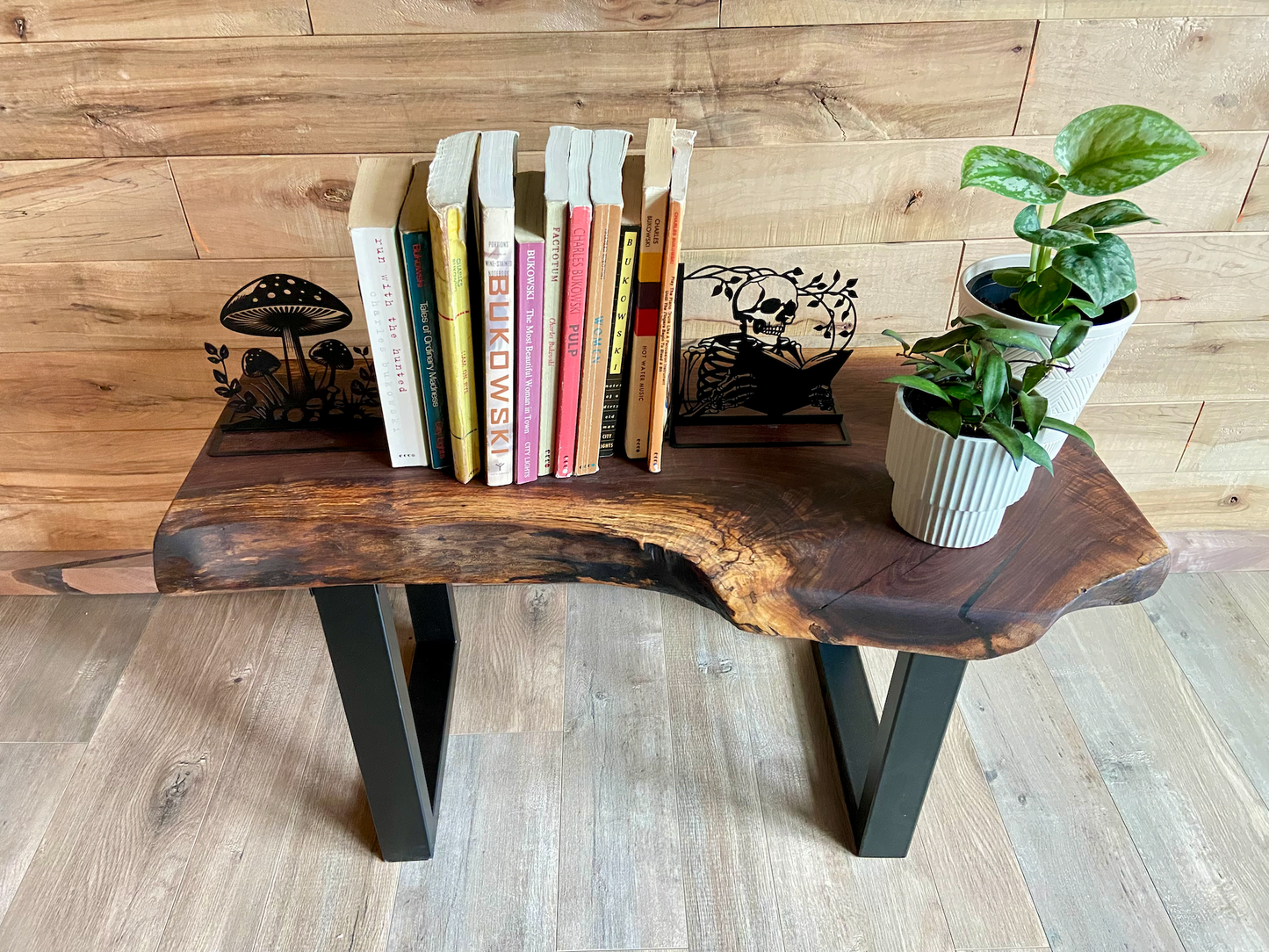 A handcrafted live edge walnut display table with black metal legs, placed against a wooden accent wall. The live edge accent table showcases natural wood grain, knots, and organic edges, adding a rustic-modern touch to the space. A live edge walnut accent table styled with a candle, notebook, pen, a book, and a potted plant. The solid wood surface has a rich, natural finish, making it a perfect accent piece for an entryway or living space.