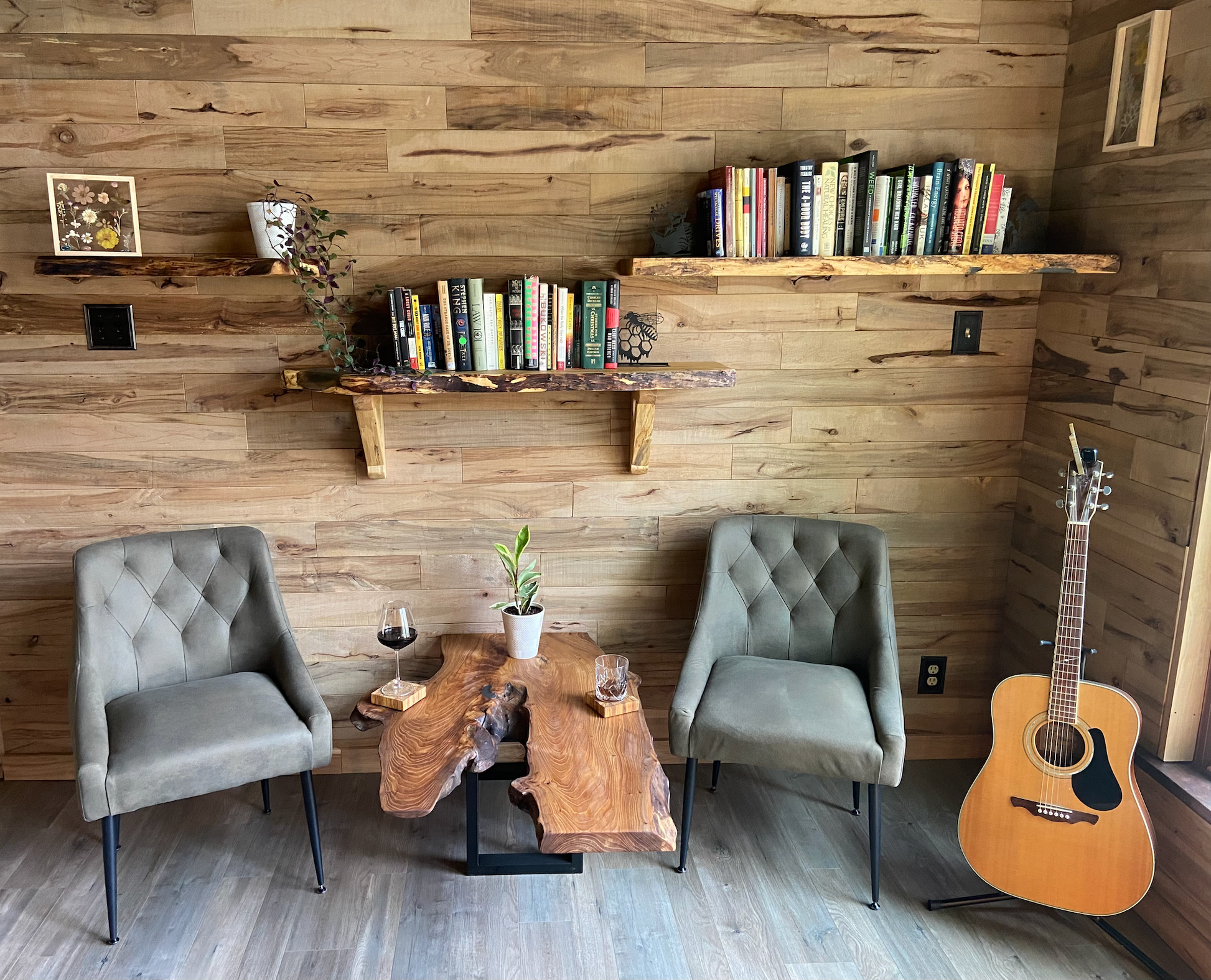Cozy room with wooden walls, two gray chairs, a wooden live edge chestnut table, and a guitar against the wall that has floating shelves on it with books.
