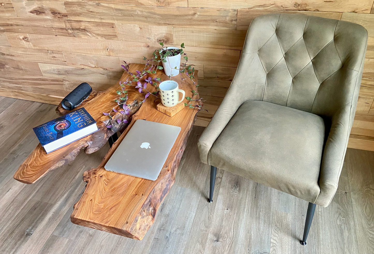 Wooden live edge table with laptop, mug, plant, and books next to a gray chair in a room with wooden floor and wall.