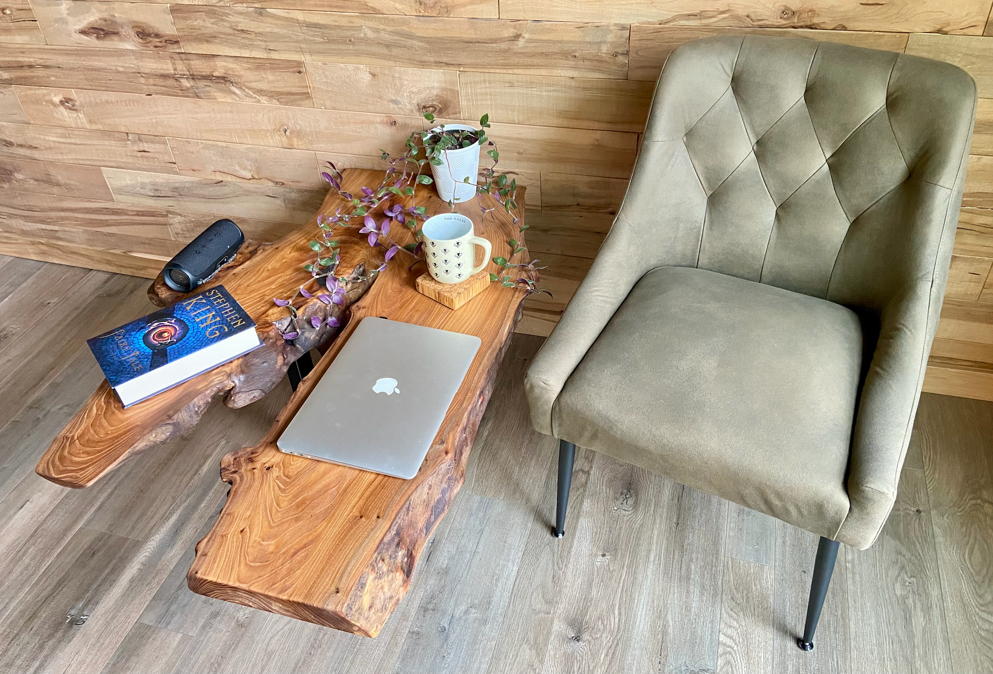 Wooden live edge table with laptop, mug, plant, and books next to a gray chair in a room with wooden floor and wall.