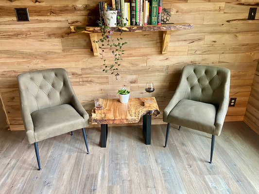 Two upholstered chairs facing a live edge wooden table with a plant and glass against a wooden wall with live edge shelves.