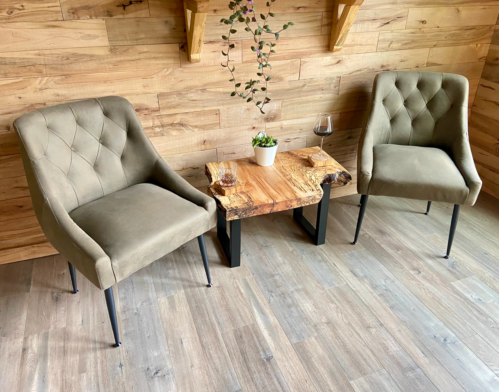 Two upholstered chairs facing a live edge wooden table with a plant and glass against a wooden wall with live edge shelves.