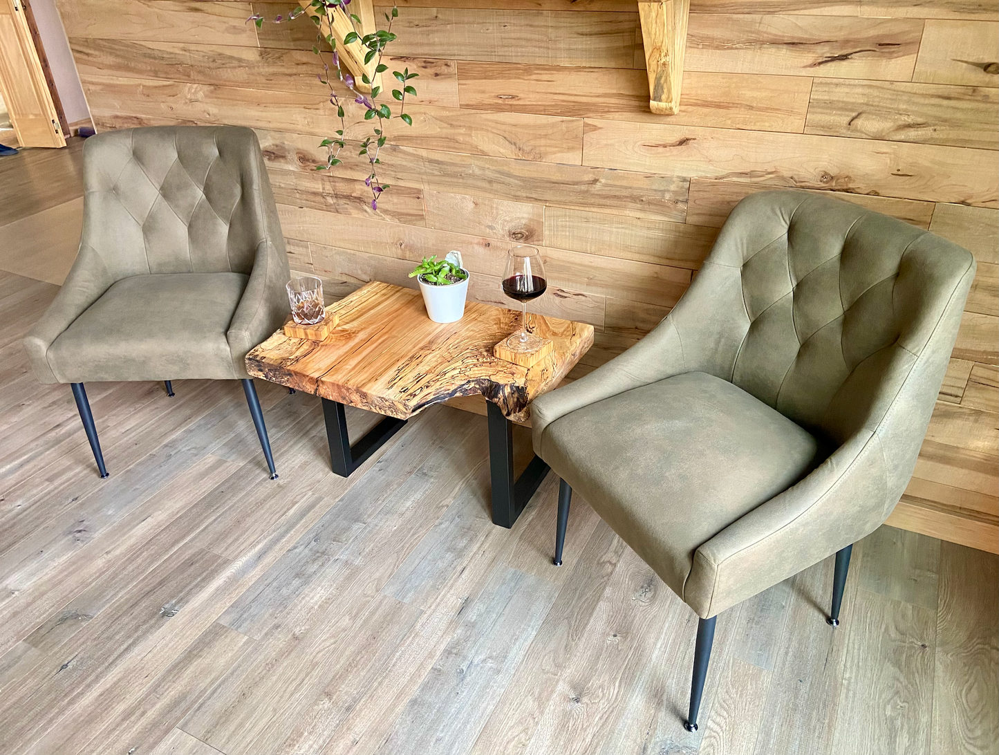 Two upholstered chairs facing a live edge wooden table with a plant and glass against a wooden wall with live edge shelves.