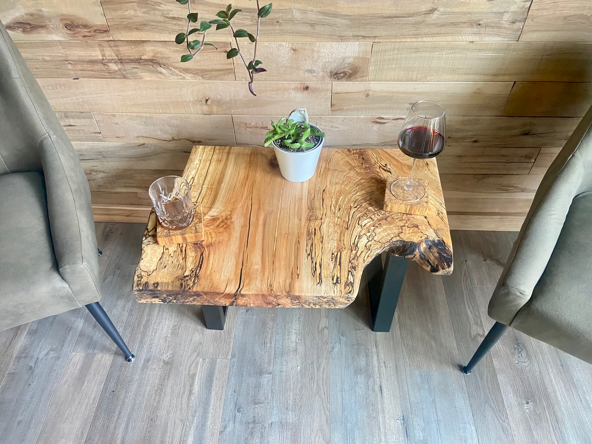 Two upholstered chairs facing a live edge wooden table with a plant and glass against a wooden wall with live edge shelves.