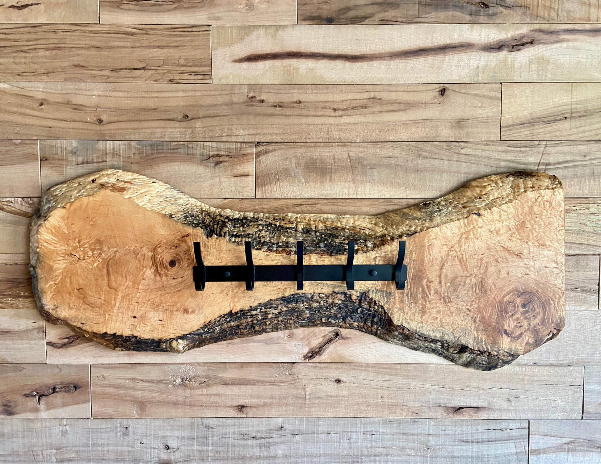 Live edge maple coat rack mounted on a wood-paneled wall, featuring 5 matte black metal hooks and natural wood grain patterns.