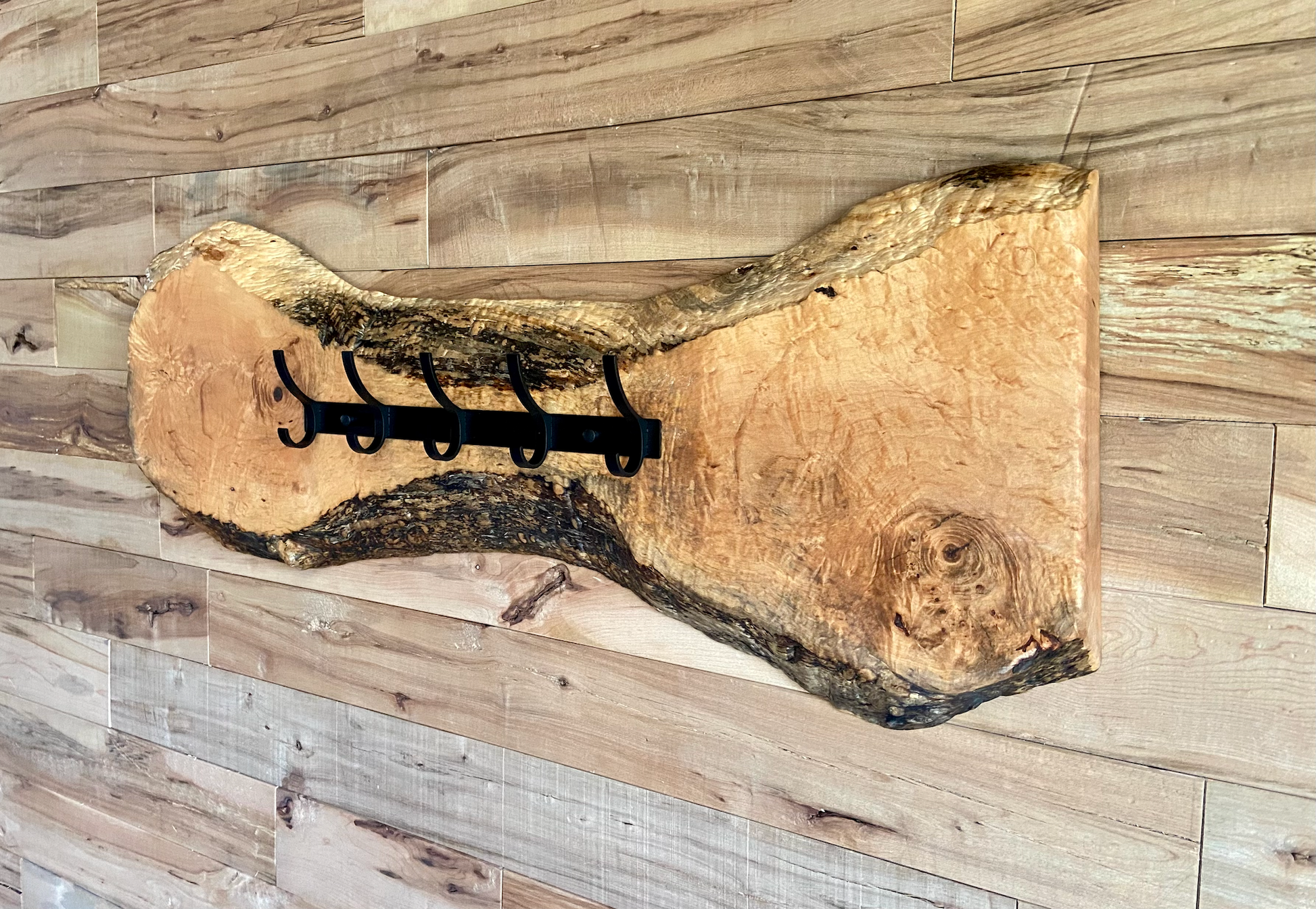 Live edge maple coat rack mounted on a wood-paneled wall, featuring 5 matte black metal hooks and natural wood grain patterns.