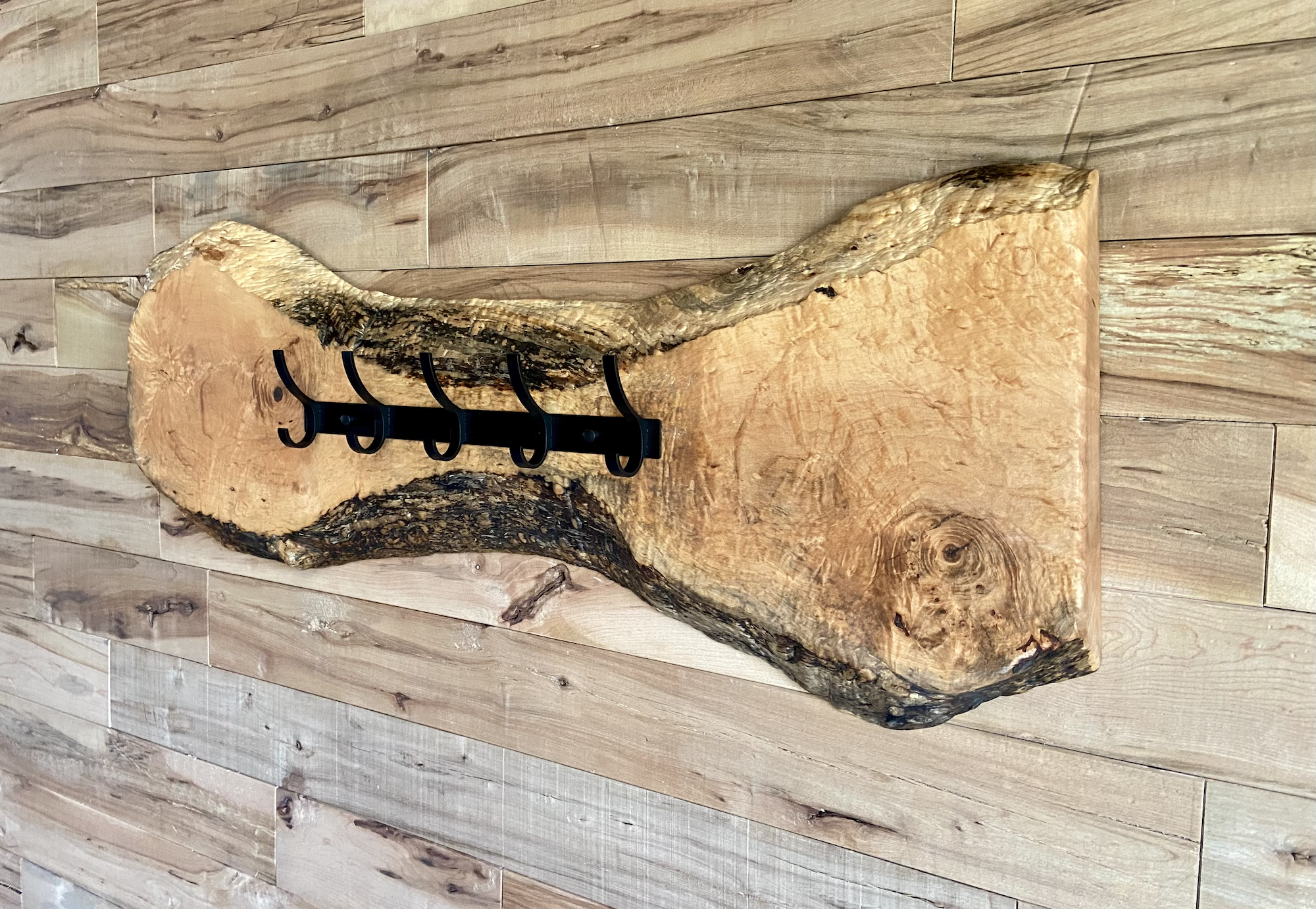 Live edge maple coat rack mounted on a wood-paneled wall, featuring 5 matte black metal hooks and natural wood grain patterns.