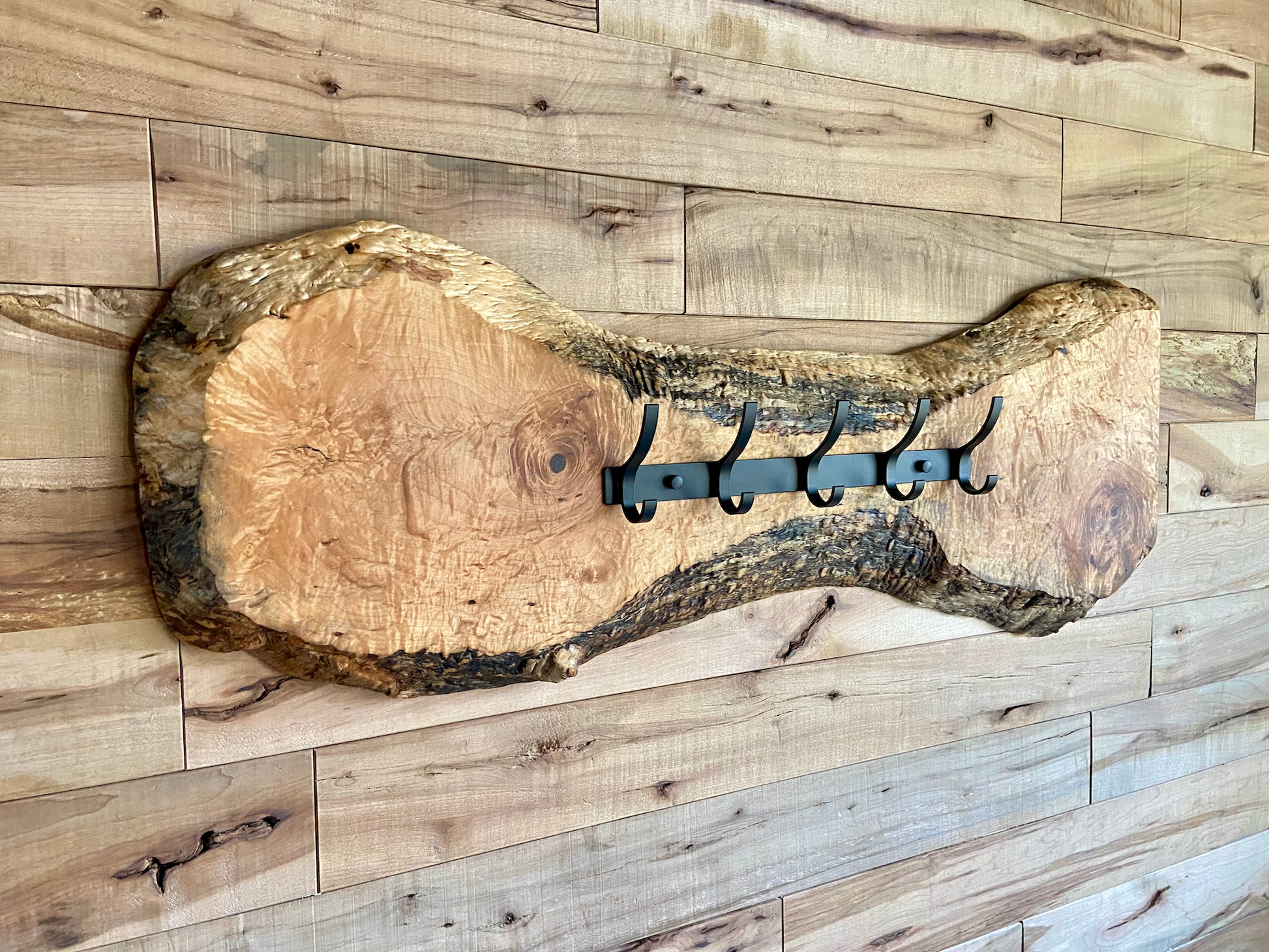 Live edge maple coat rack mounted on a wood-paneled wall, featuring 5 matte black metal hooks and natural wood grain patterns.
